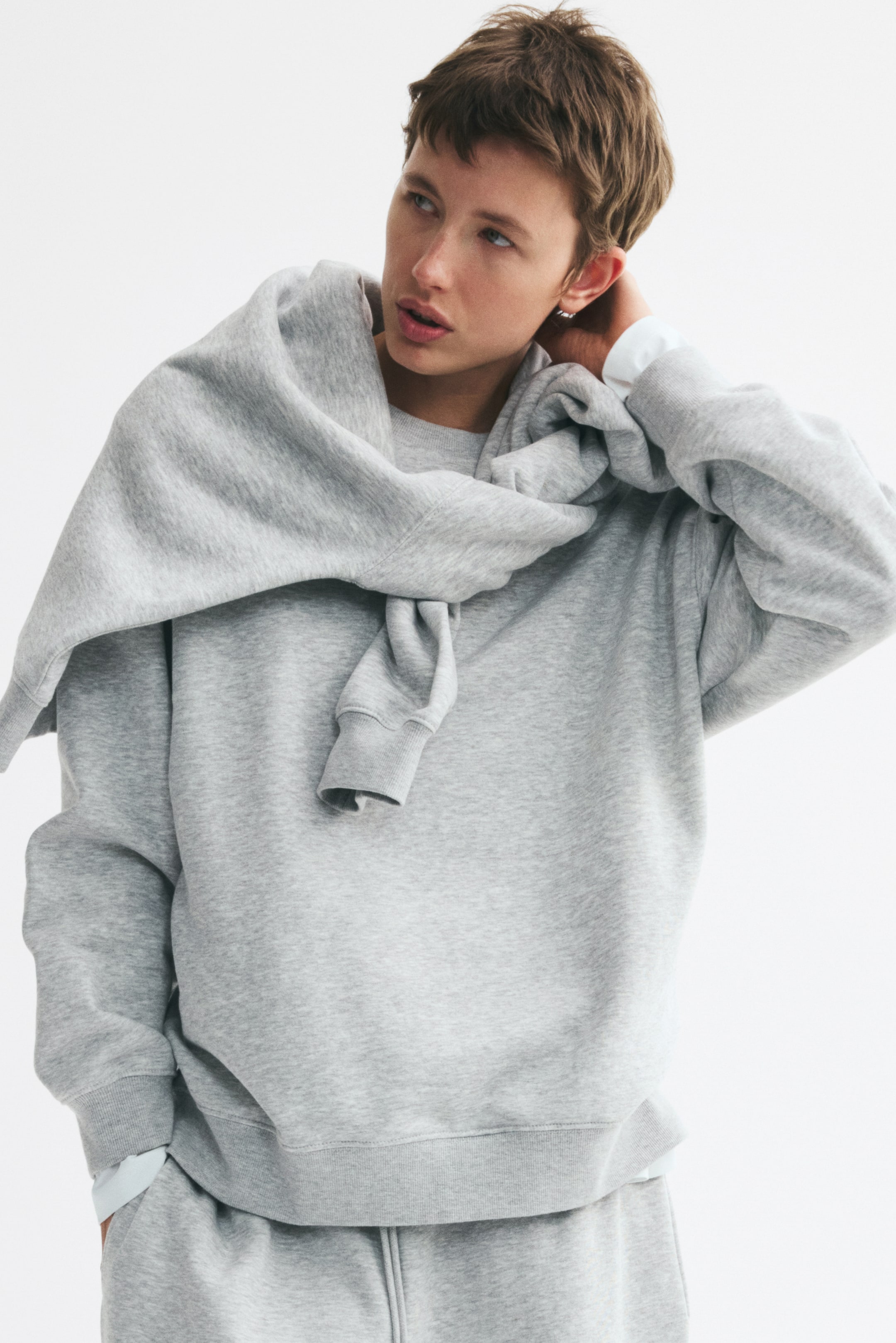 Näytä suurempi kuva: Vaaleanharmaa oversized-collegepaita, jossa on pyöreä ribbineuloksella viimeistelty pääntie, laskettu olkasauma, pitkät hihat sekä ribbiresorit hihansuissa ja helmassa, on mallin yllä. Toinen samanlainen collegepaita on asetettu mallin hartioille, ja alla on kerroksittain valkoinen pitkähihainen paita.