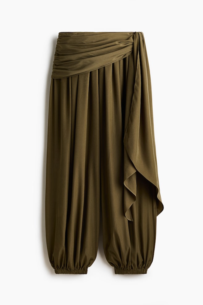 Pantalonii țesuți de culoare verde măsliniu sunt prezentați din față, arătând o ușoară strălucire și o talie înaltă cu elastic acoperit. O secțiune suprapusă se leagă pe partea dreaptă, creând drapaje în față, iar picioarele balon se adună într-un elastic îngust la tivuri. - 1