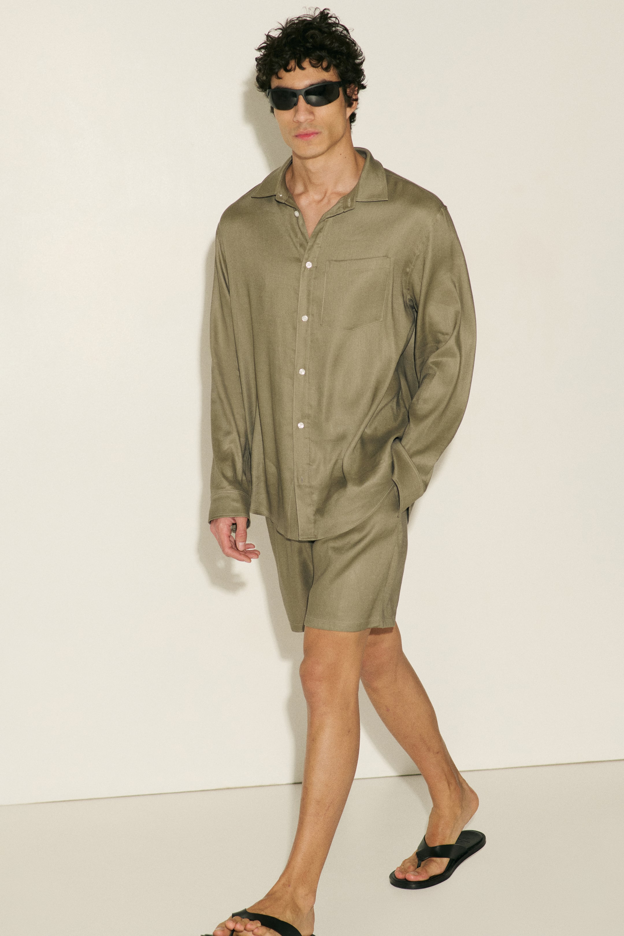 Agrandir l'image: Un ensemble assorti de chemise à manches longues et de short vert olive est présenté sous un léger angle, avec un col à revers, une fermeture boutonnée à l'avant avec des boutons blancs et une poche poitrine plaquée unique sur la chemise, tandis que le short à coupe décontractée s'étend jusqu'au milieu de la cuisse.
