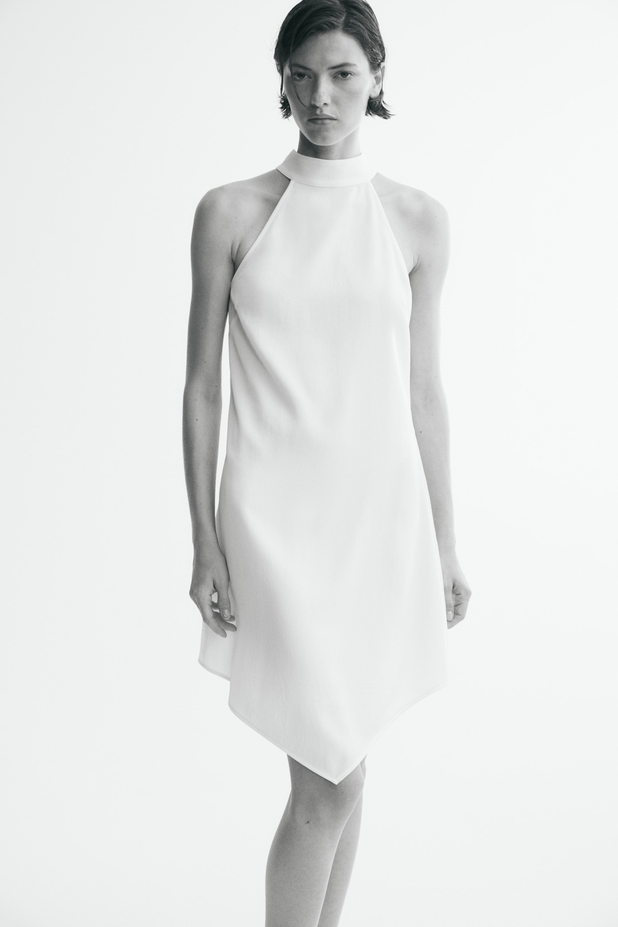 Agrandir l'image: Une robe courte blanche avec un haut ajusté, un col montant et un ourlet asymétrique en forme de mouchoir à l'avant est portée par le mannequin.