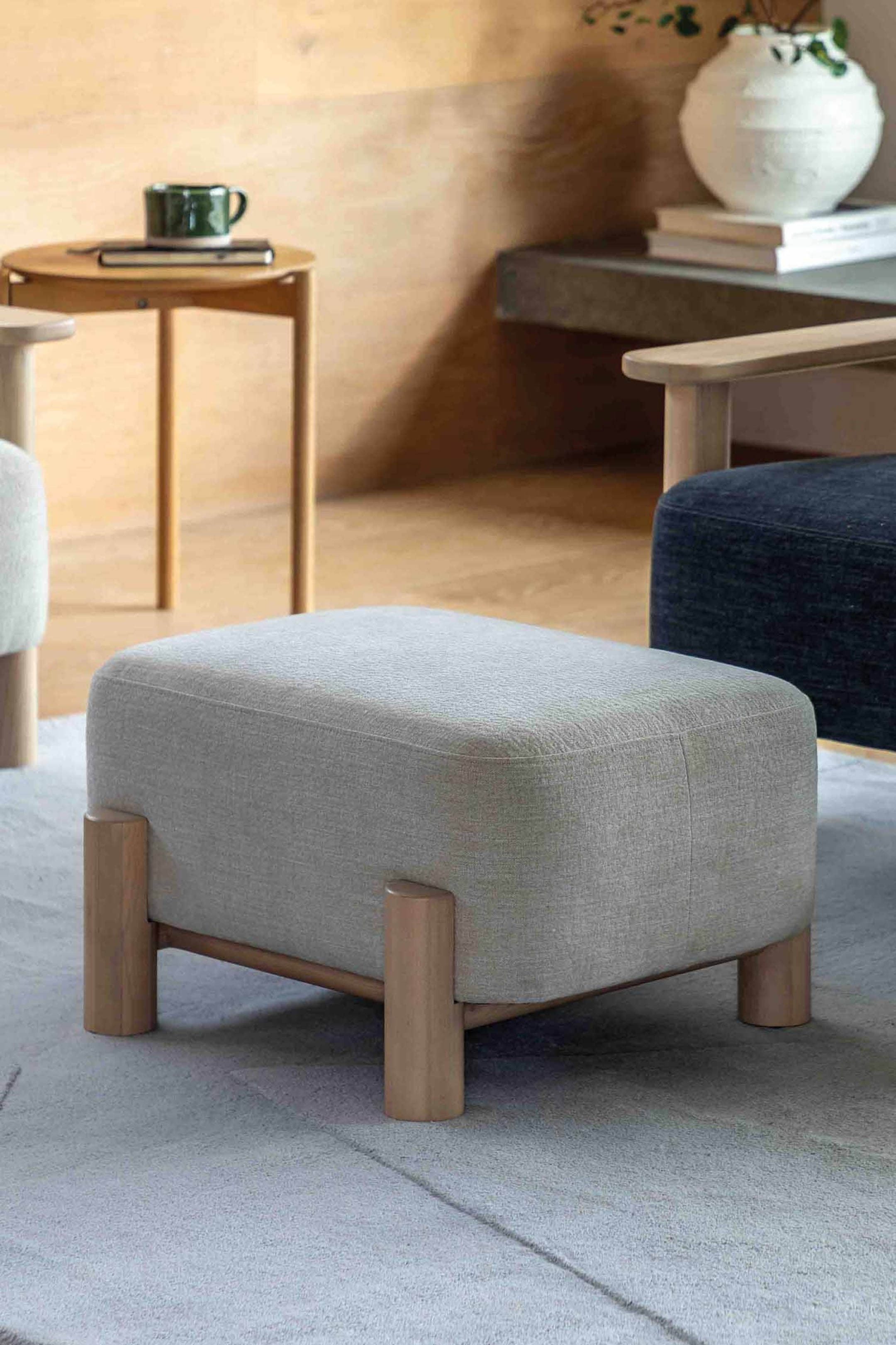 View larger image: The ottoman is upholstered in a light grey textured fabric, showcasing a rectangular shape with rounded top edges. It is supported by four cylindrical light wood legs connected by a wooden frame, positioned at a slight angle towards the right.
