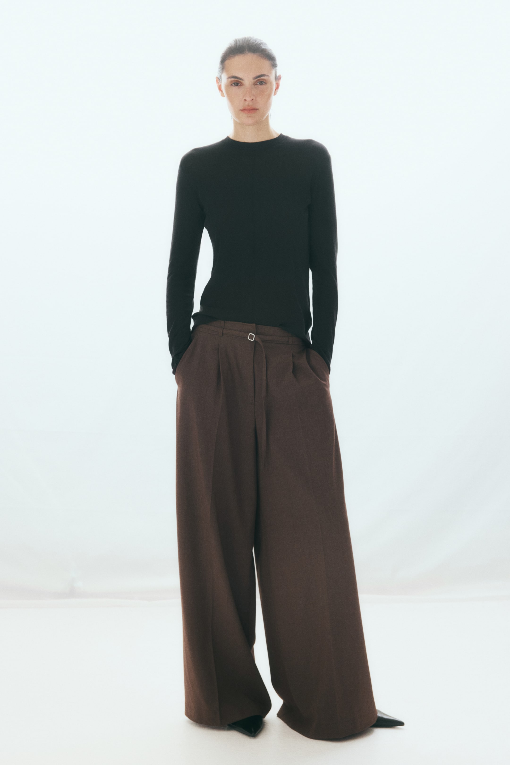 Voir une image plus grande: Pantalon tailleur brun à jambes larges, doté d'une taille haute, d'une ceinture étroite ajustable et de plis repassés, porté par le mannequin. Le mannequin est debout, les mains insérées dans les poches latérales en biais, et agence le pantalon avec un haut noir à manches longues et des chaussures à bout pointu.