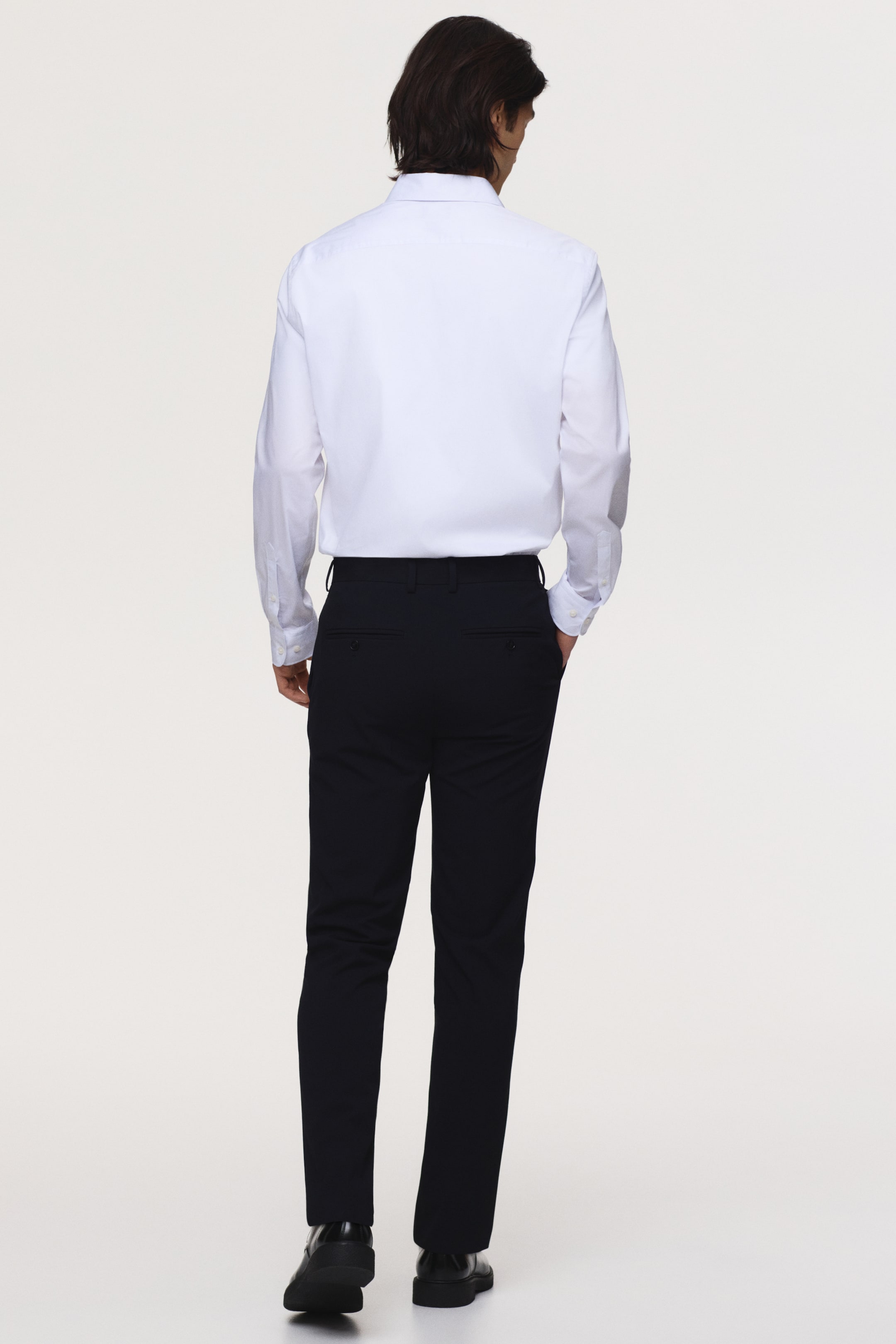 Agrandir l'image: Pantalon tailleur foncé, avec une coupe ajustée et des poches arrière passepoilées avec boutons, porté par le mannequin qui place ses mains dans les poches latérales.