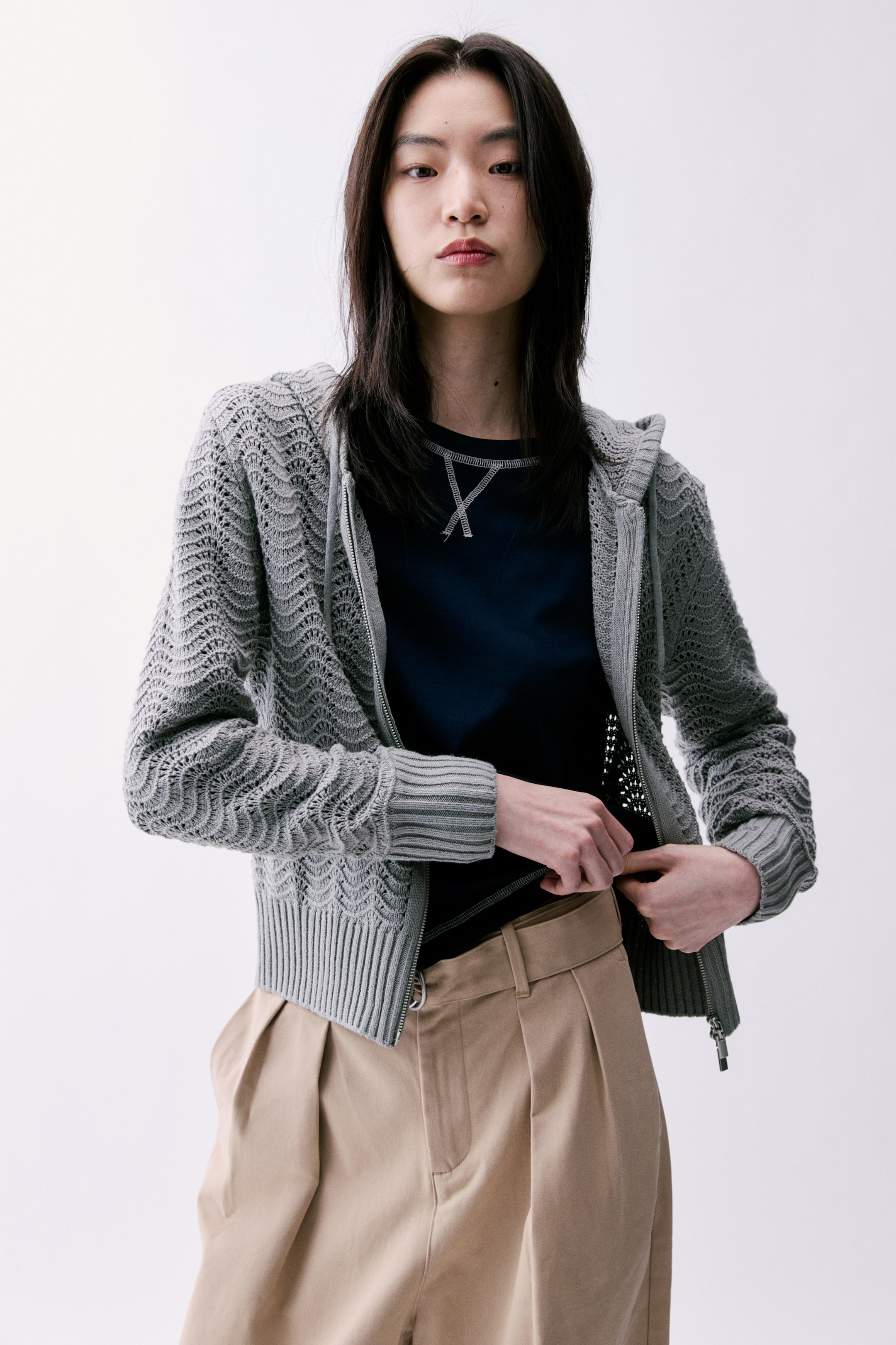 拡大画像を表示: グレーのニットフード付きカーディガンは、ボディと袖に波状のオープンニットパターンが特徴です。フルレングスのジッパー、リブ編みのカフス、リブ編みの裾を備えており、ダークなTシャツの上にオープンに着用され、そのクロップドフィットを引き立てています。
