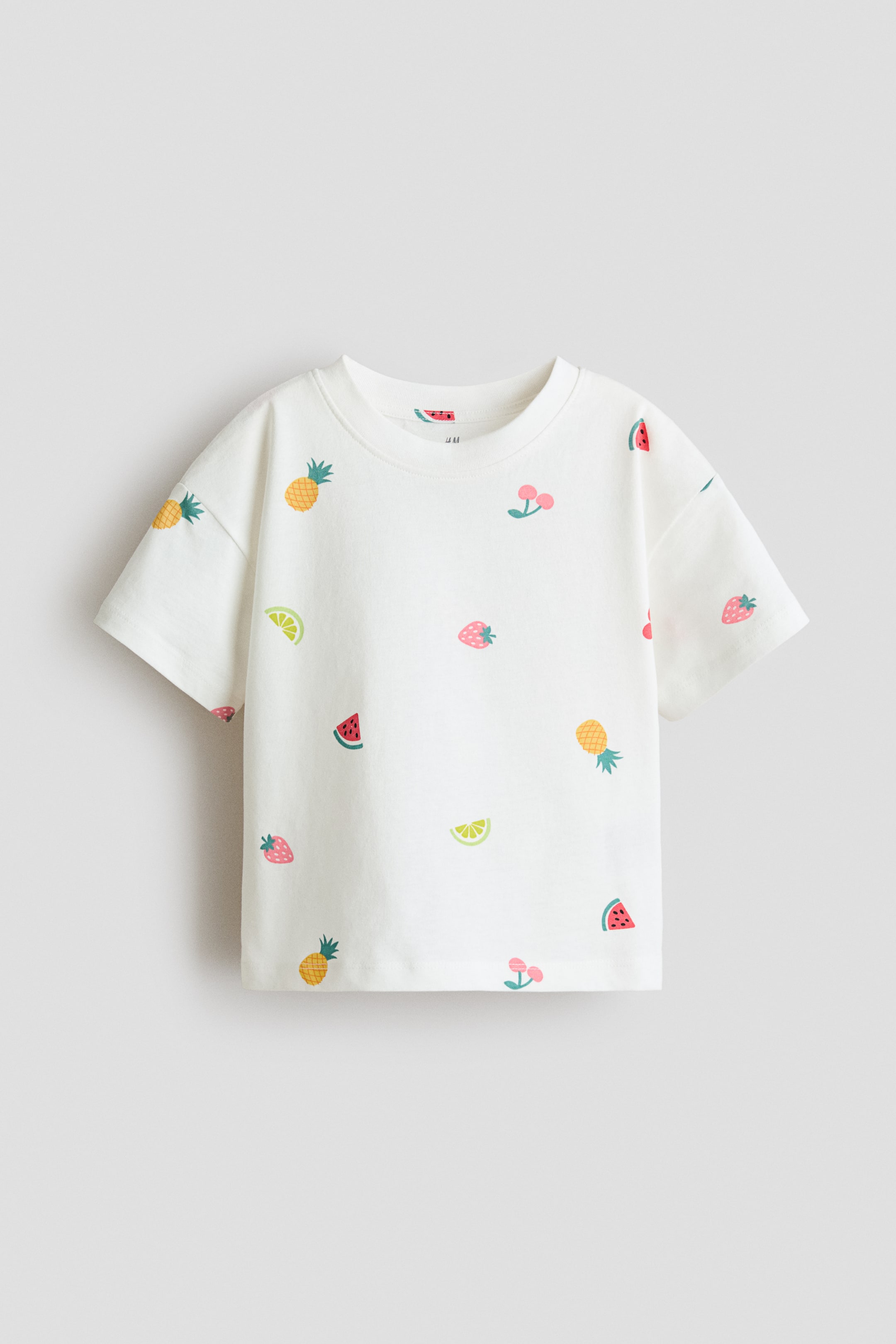 Größeres Bild ansehen: Ein weißes T-Shirt wird von vorne präsentiert, mit einem gerippten Rundhalsausschnitt und kurzen Ärmeln. Es zeigt ein verstreutes Muster aus bunten Fruchtmotiven, darunter Ananas, Kirschen, Wassermelonenstücke, Erdbeeren und Limettenscheiben, über den gesamten Stoff verteilt.