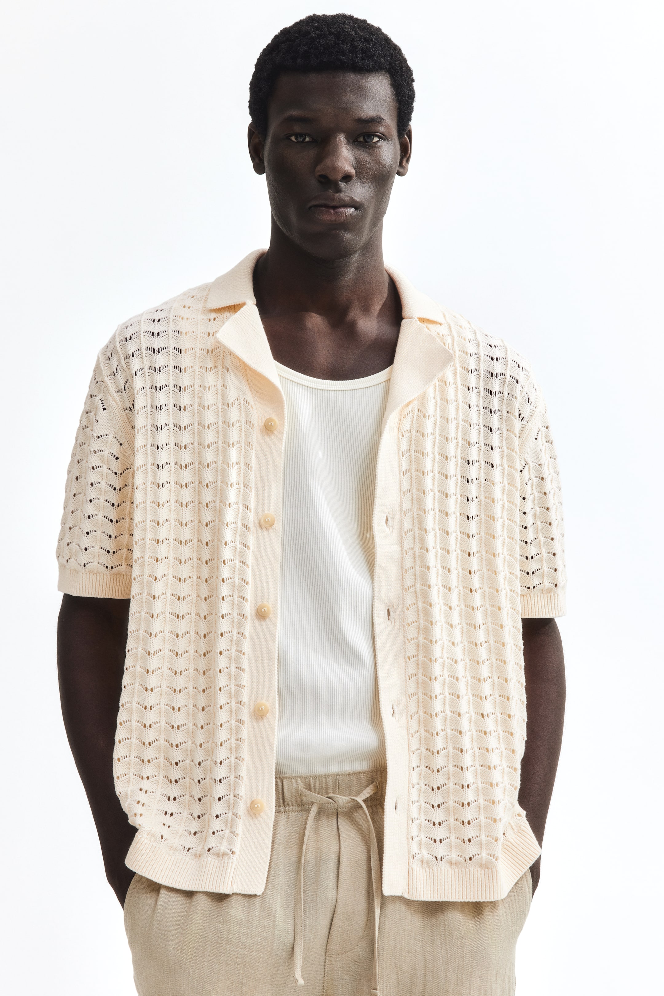 View larger image: Presenting a cream open-knit short-sleeve shirt showcasing a chevron pattern, camp collar, and ribbed cuffs and hem. Styled unbuttoned over a white ribbed tank top.