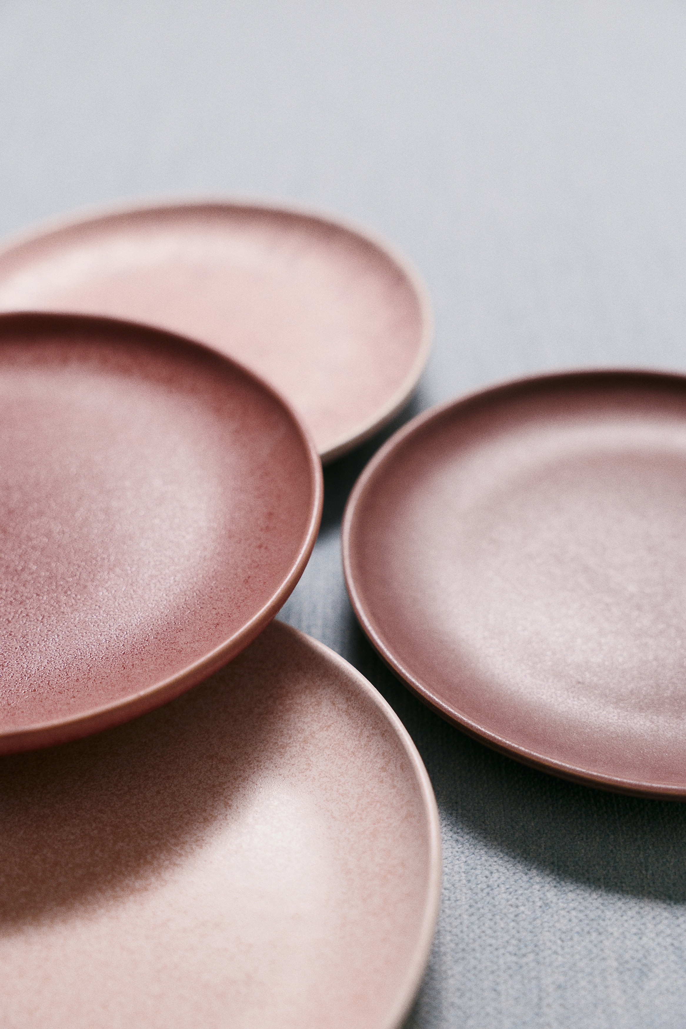 Four round stoneware side plates are displayed in a top-down view, each featuring unique glazed textures and shades of pink, mauve, and plum. The plates showcase different speckled, mottled, and smooth matte finishes across their circular designs.
