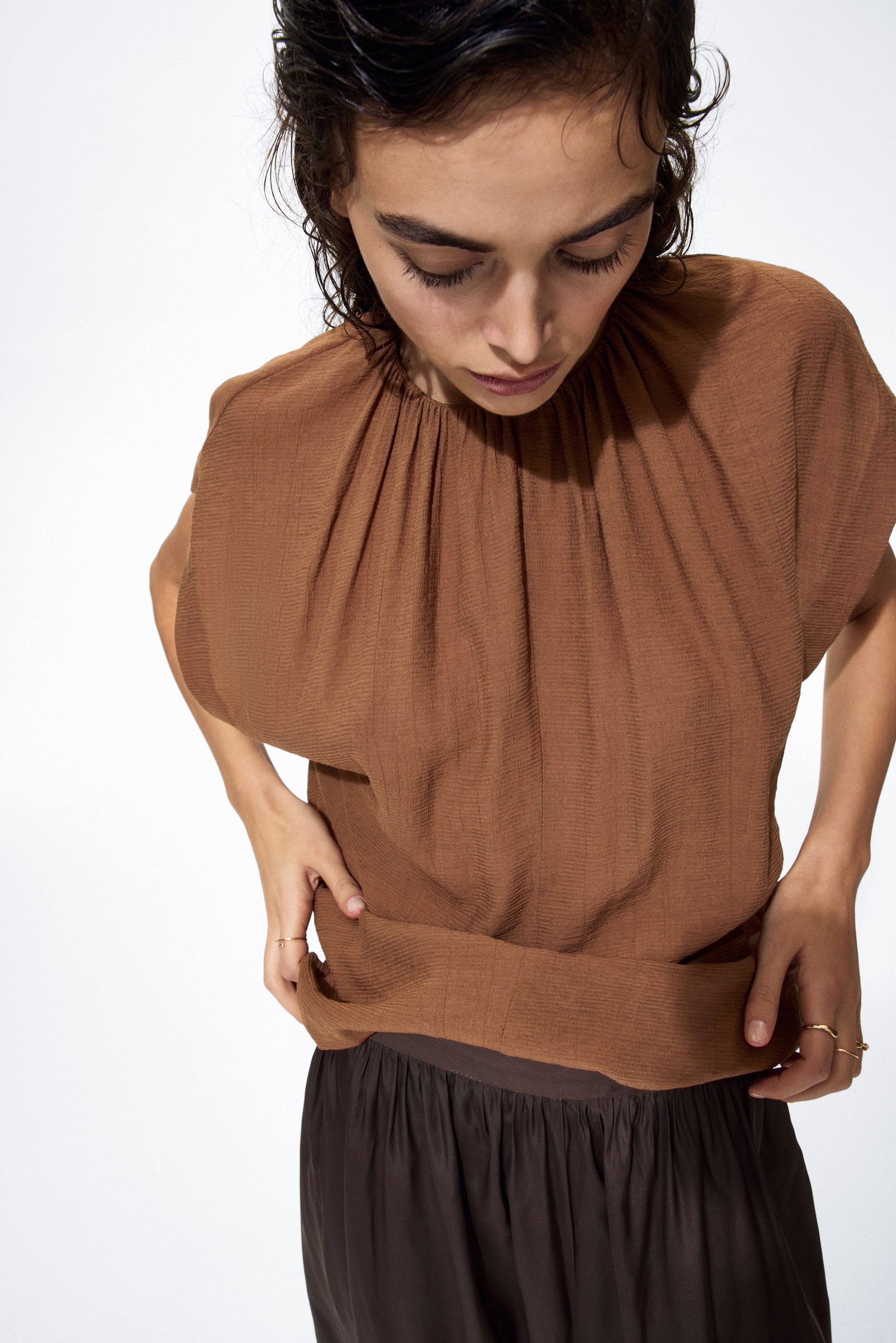 View larger image: Presenting a brown crinkled top, adorned with a gathered round neckline and short, wide sleeves. The loose hem is stylishly tucked into dark brown trousers, highlighting the top's relaxed fit.