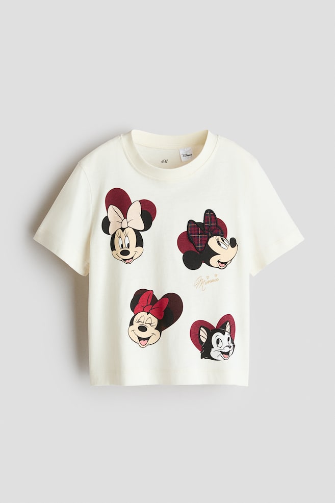 A cream-coloured, short-sleeved cotton jersey T-shirt is shown from the front, featuring a round, rib-trimmed neckline. The tee displays printed graphics of Minnie Mouse in three different poses and the character Figaro, each set against a dark red heart backdrop. - 1