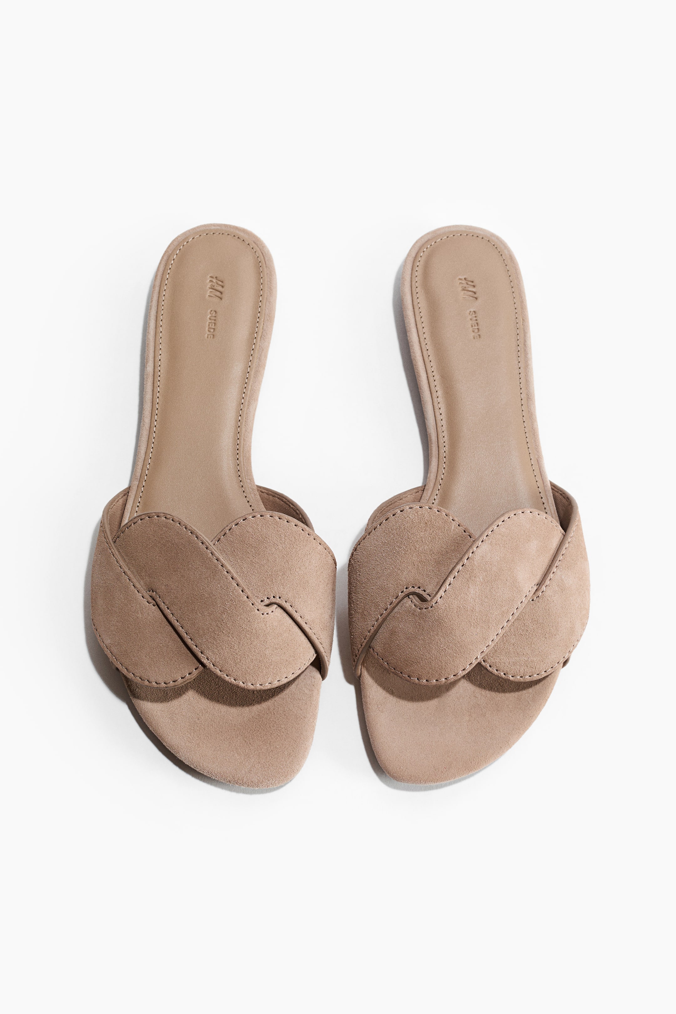 הצגת תמונה גדולה יותר: זוג סנדלי סלייד עשויים זמש בצבע חום בהיר מוצגים מלפנים. כל סנדל כולל עיצוב רצועה קדמית שזורה עם תפרים גלויים לאורך הקצוות, והסוליות הפנימיות מציגות את הכיתוב "H&M SUEDE".
