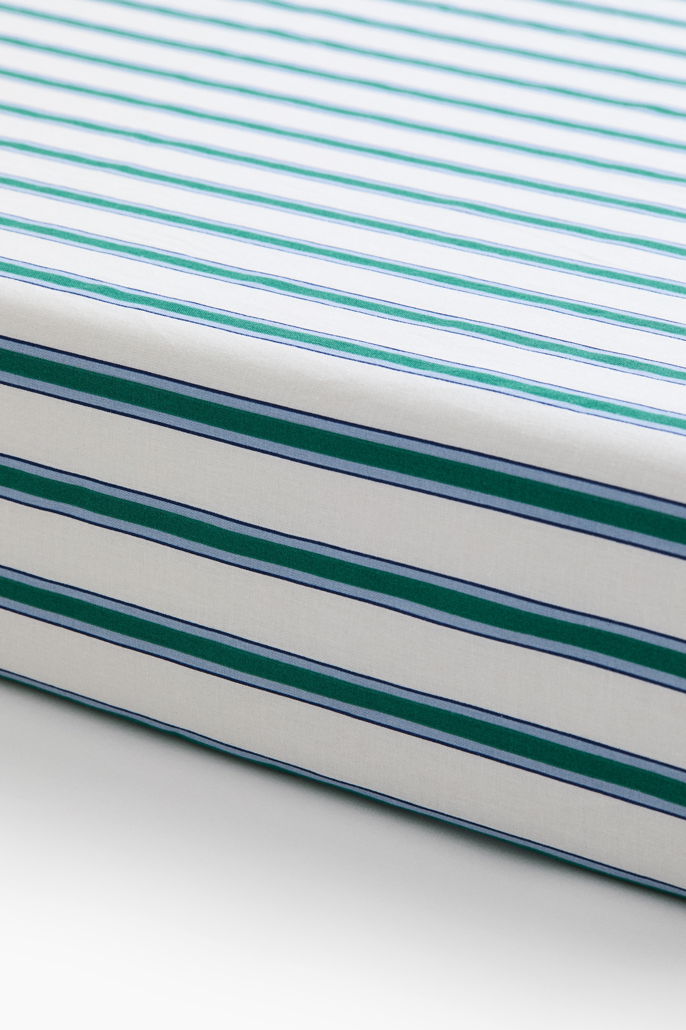 Un drap-housse en tissage à motifs présente une série de rayures vert foncé, fines et épaisses, sur un fond blanc. Un oreiller vert orné de divers emblèmes graphiques repose sur la surface du drap.