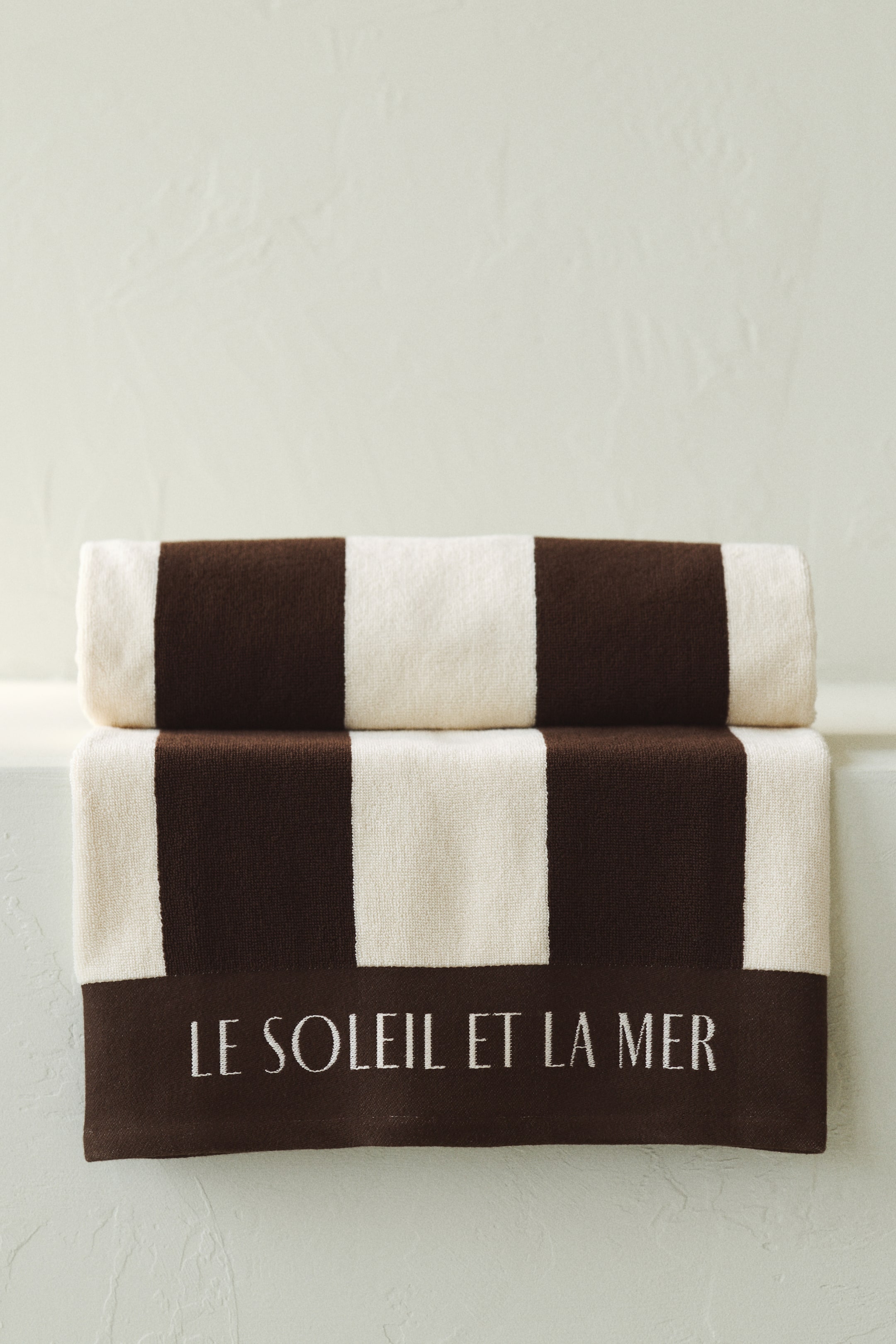 Grotere afbeelding bekijken: Twee handdoeken zijn gestapeld, met afwisselende horizontale strepen in bruin en gebroken wit. De bovenste handdoek is opgerold, terwijl de onderste handdoek plat is gevouwen en de tekst "LE SOLEIL ET LA MER" geborduurd op het onderste bruine gedeelte toont.