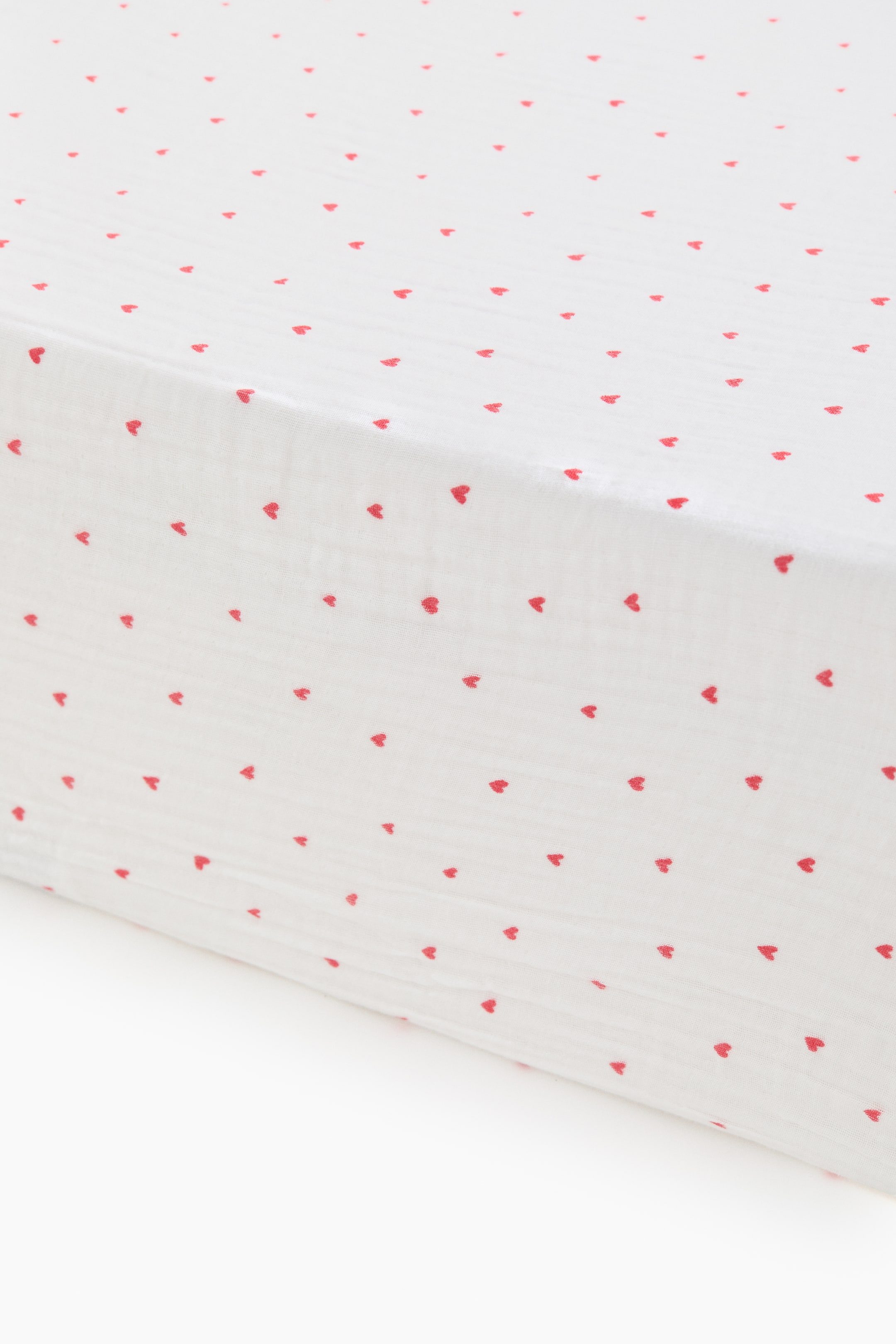 View larger image: A fitted crib sheet shows its soft cotton muslin texture. It features an all-over pattern of small red hearts on a white background, with its fitted shape curving along an edge.