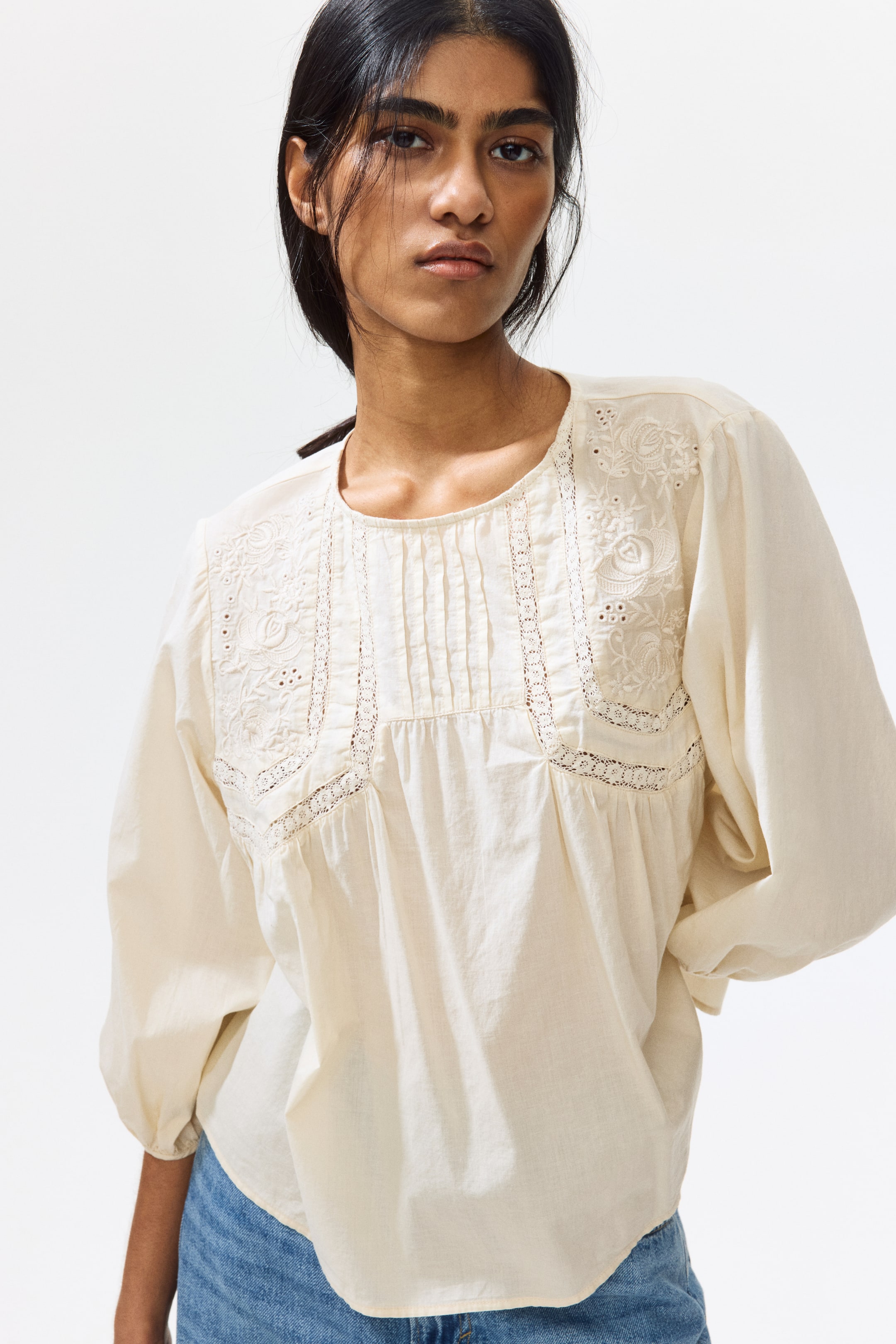 View larger image: A cream-coloured blouse is showcased front-facing, highlighting a round neckline with pintucks and lace inserts on the front yoke. Floral embroidery adorns the upper chest and shoulders, while the long sleeves appear voluminous.