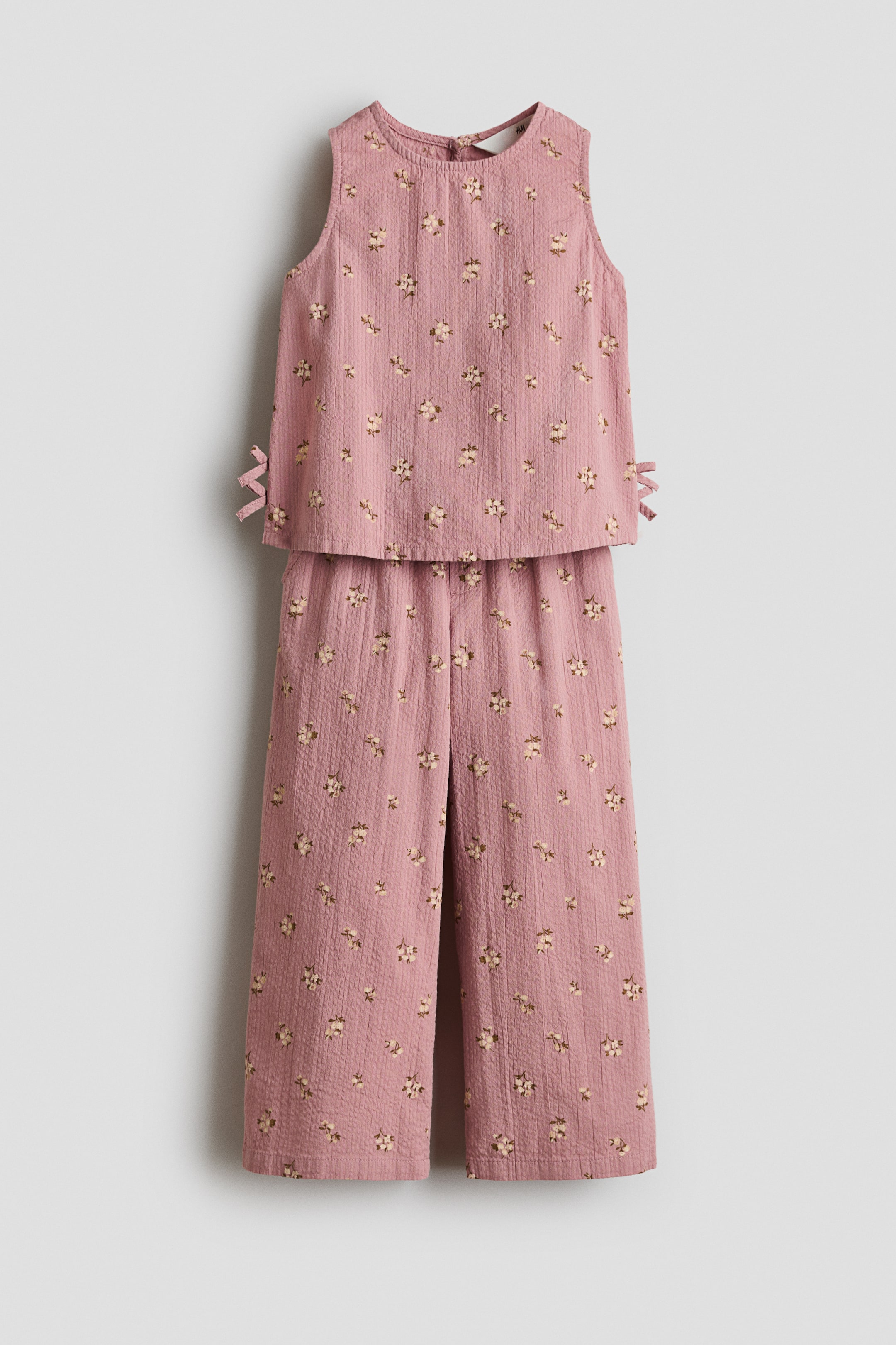 View larger image: A two-piece seersucker set in dusty pink with a small floral pattern is shown from the front. The sleeveless top features a round neckline and small bow details on the sides of the hem, while the matching pants display a straight-leg cut.