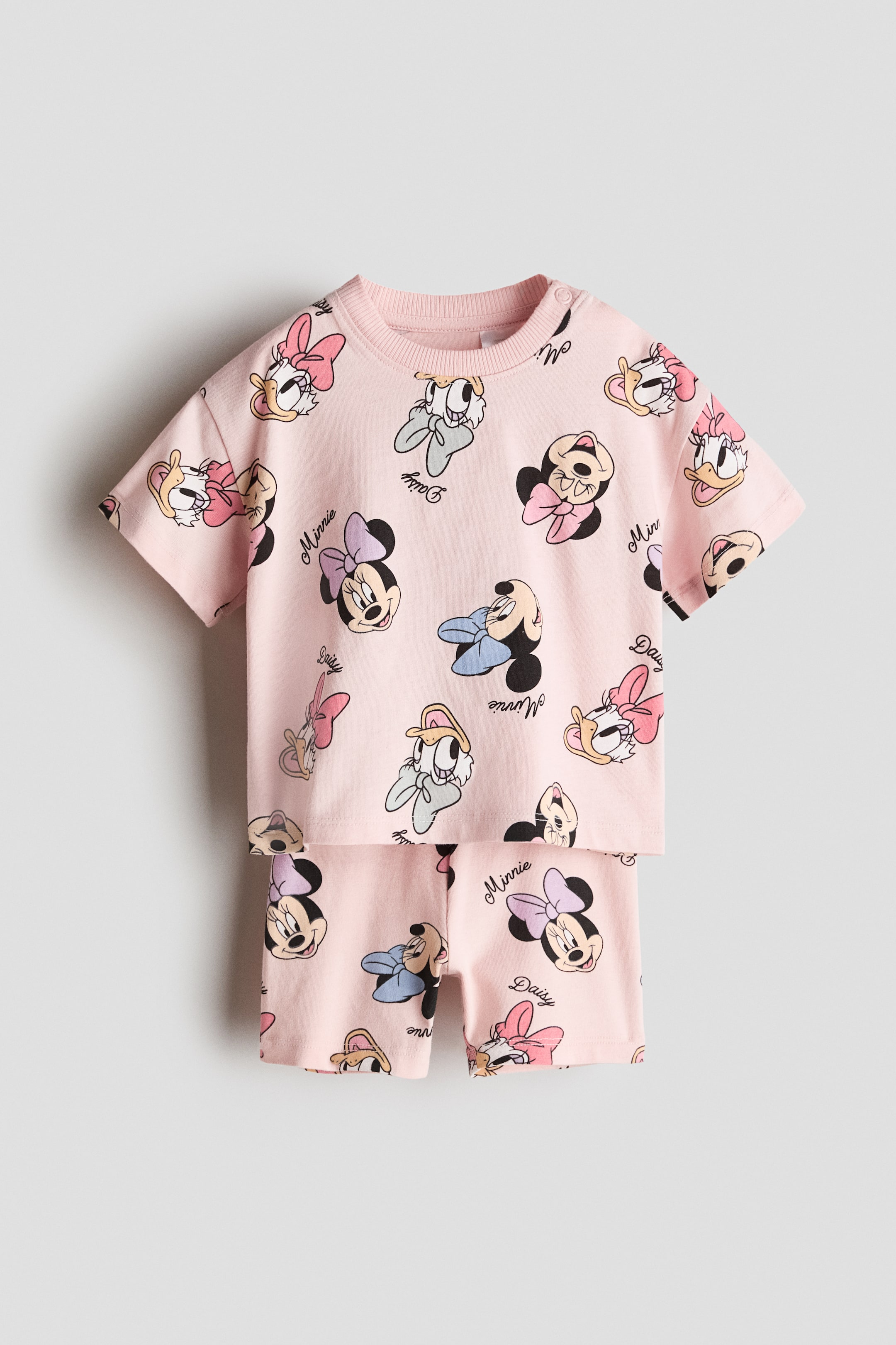 View larger image: A 2-piece pink cotton set is showcased from the front, featuring a short-sleeved t-shirt with a ribbed crew neck and snap buttons on the left shoulder, accompanied by matching shorts. Both pieces are adorned with an all-over print of Minnie Mouse and Daisy Duck character illustrations.