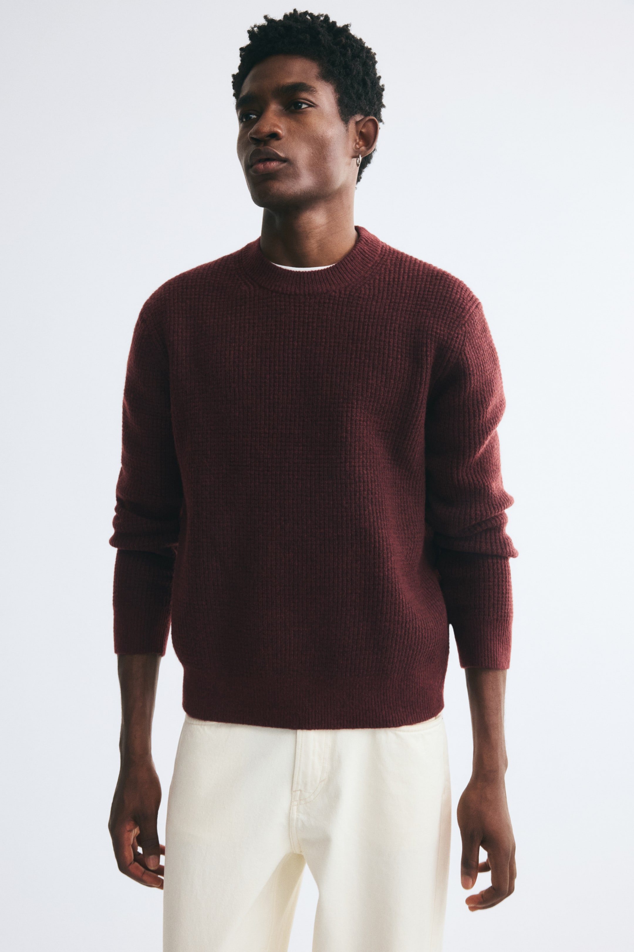 View larger image: The model is wearing a burgundy textured knit sweater featuring a round ribbed neckline, ribbed cuffs, and a ribbed hem. The relaxed-fit sweater is layered over a white collar and paired with off-white trousers.