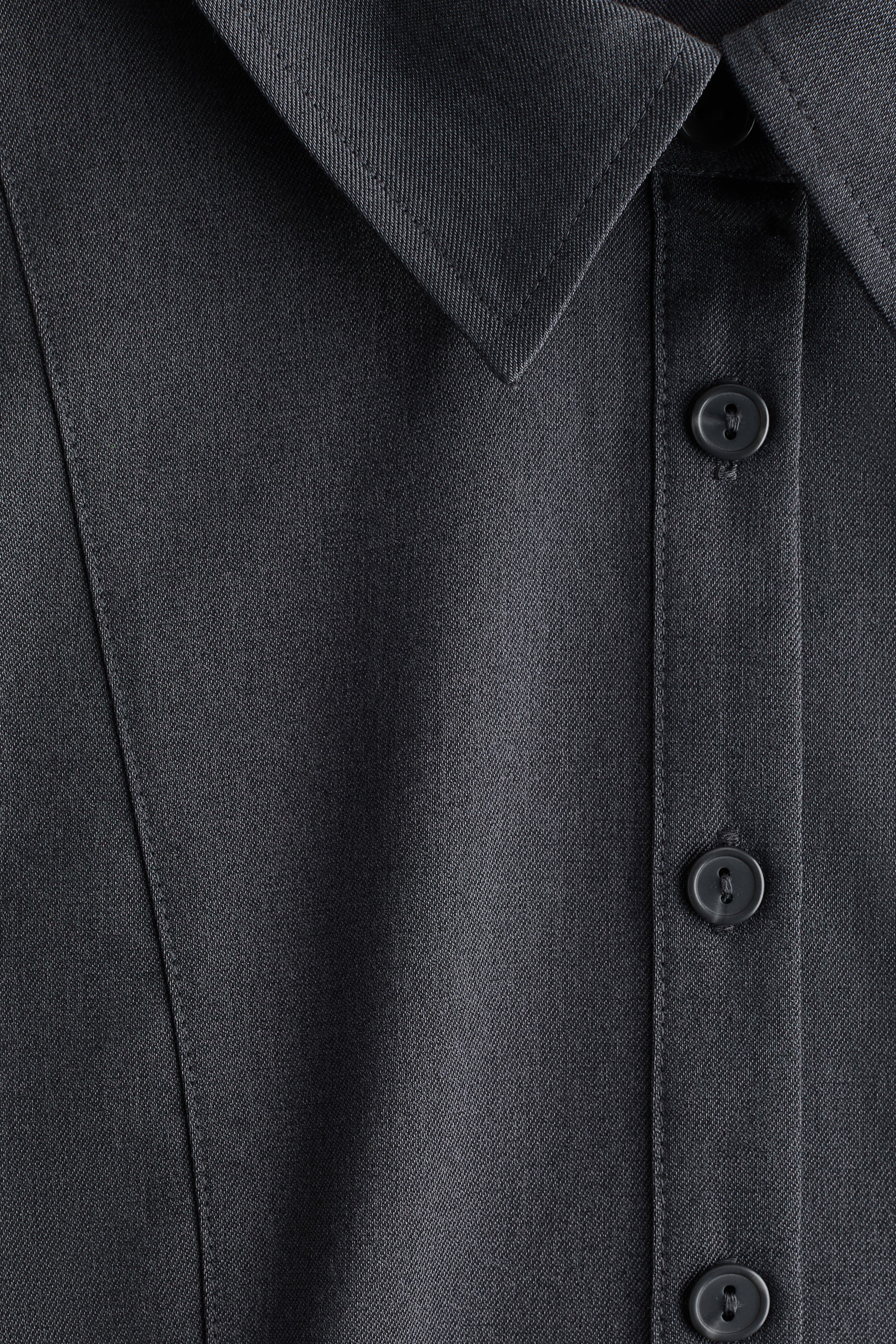拡大画像を表示: 暗いグレーのツイル衣服には、クラシックなポイントカラーとフロントボタンのプラケットが特徴です。同じく暗いグレーのボタンが3つ、プラケットを留め、左側の生地には縦の縫い目のディテールが見えます。
