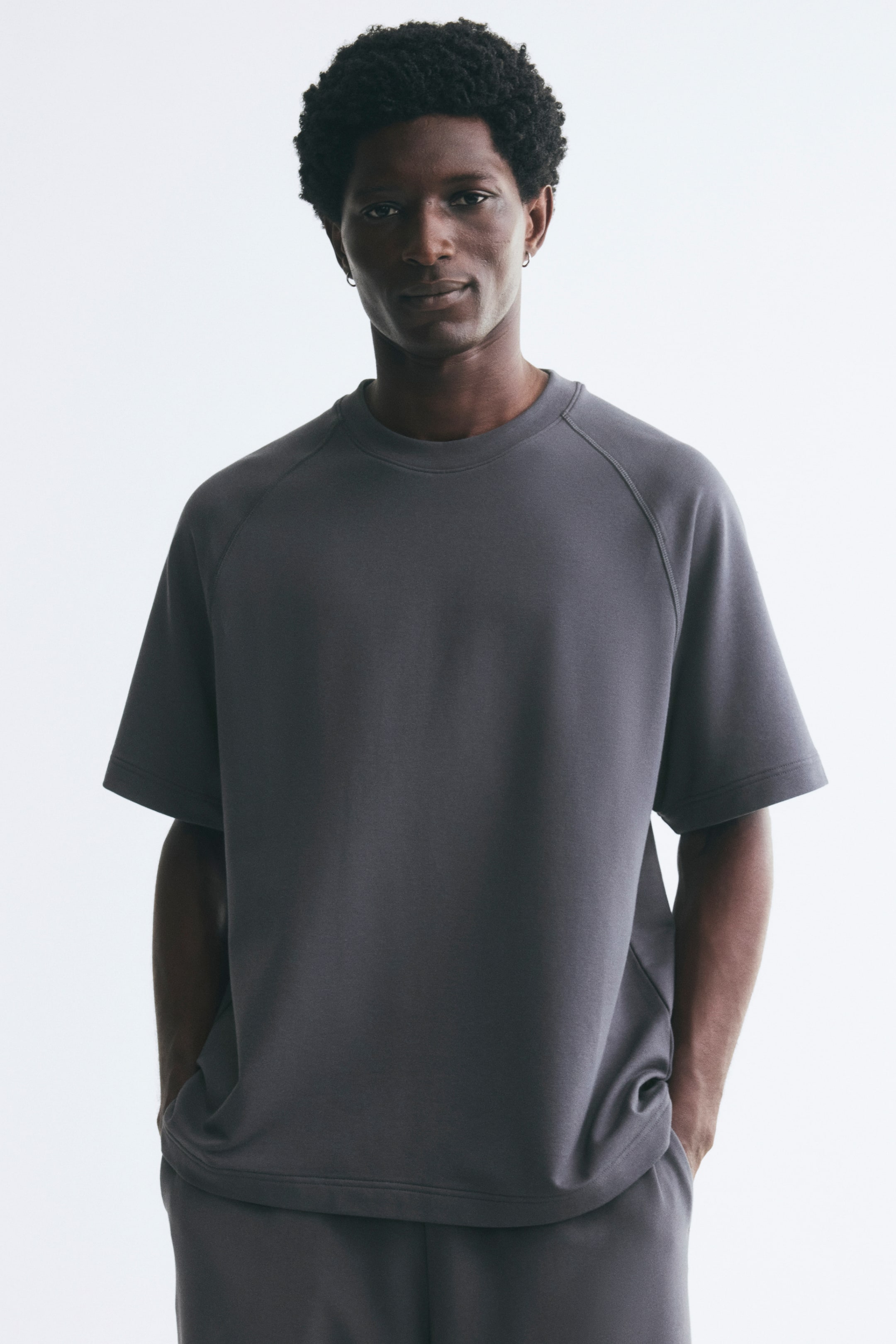 Voir une image plus grande: Un t-shirt en jersey épais gris foncé, à coupe décontractée, doté d’un col rond à bord-côte et de manches raglan courtes, est porté par le mannequin. Le t-shirt est agencé à un bas assorti gris foncé.
