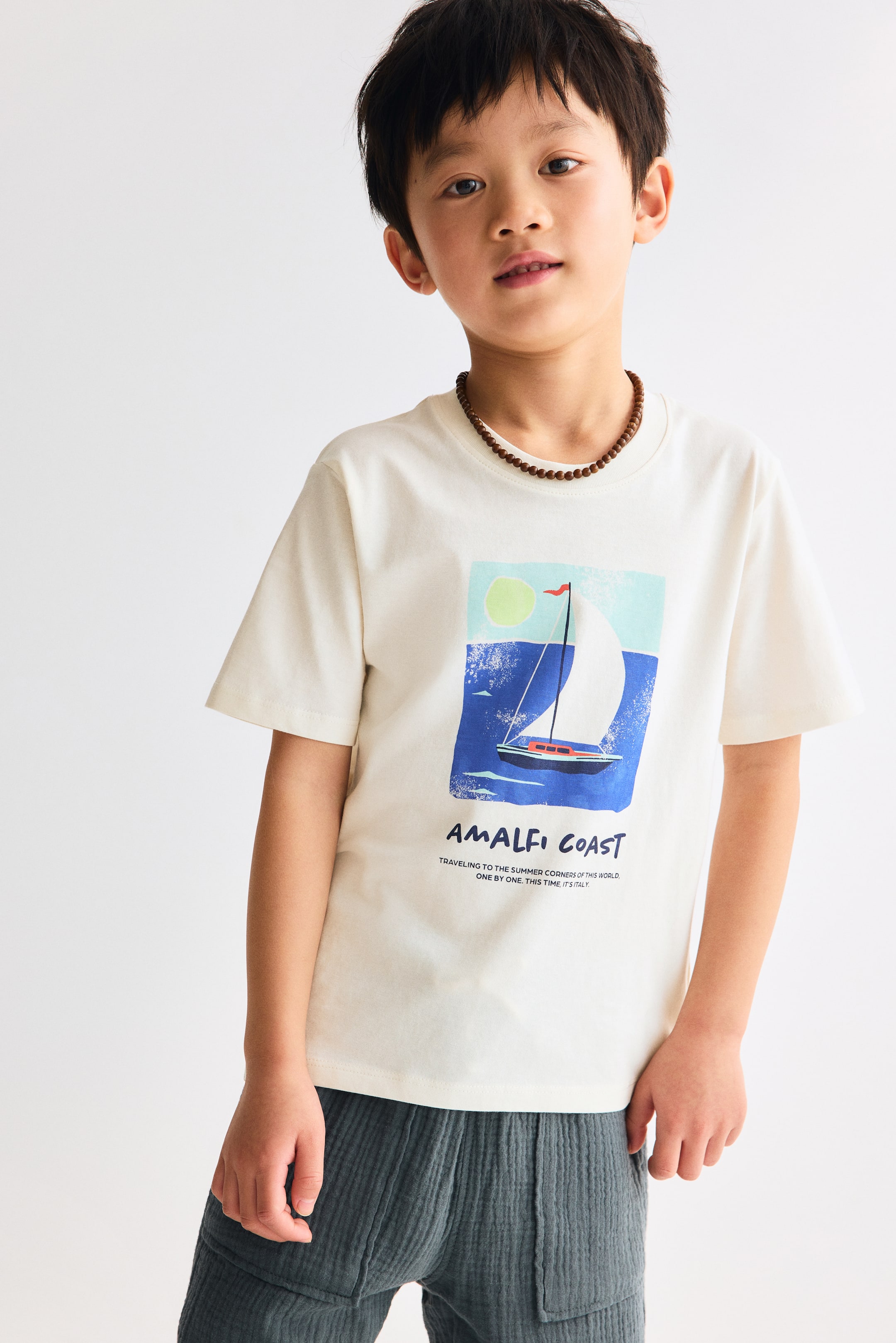 Agrandir l'image: Le t-shirt à manches courtes blanc cassé, vu de face, présente un col rond et un imprimé graphique rectangulaire sur le devant. L'imprimé représente un voilier sur une eau bleue avec un soleil vert clair et un ciel bleu clair, accompagné du texte "CÔTE AMALFITAINE" et d'un texte plus petit "VOYAGE VERS LES COINS D'ÉTÉ DE CE MONDE UN PAR UN. CETTE FOIS, C'EST L'ITALIE" positionné en dessous. Un collier de perles marron repose sur l'encolure du t-shirt.