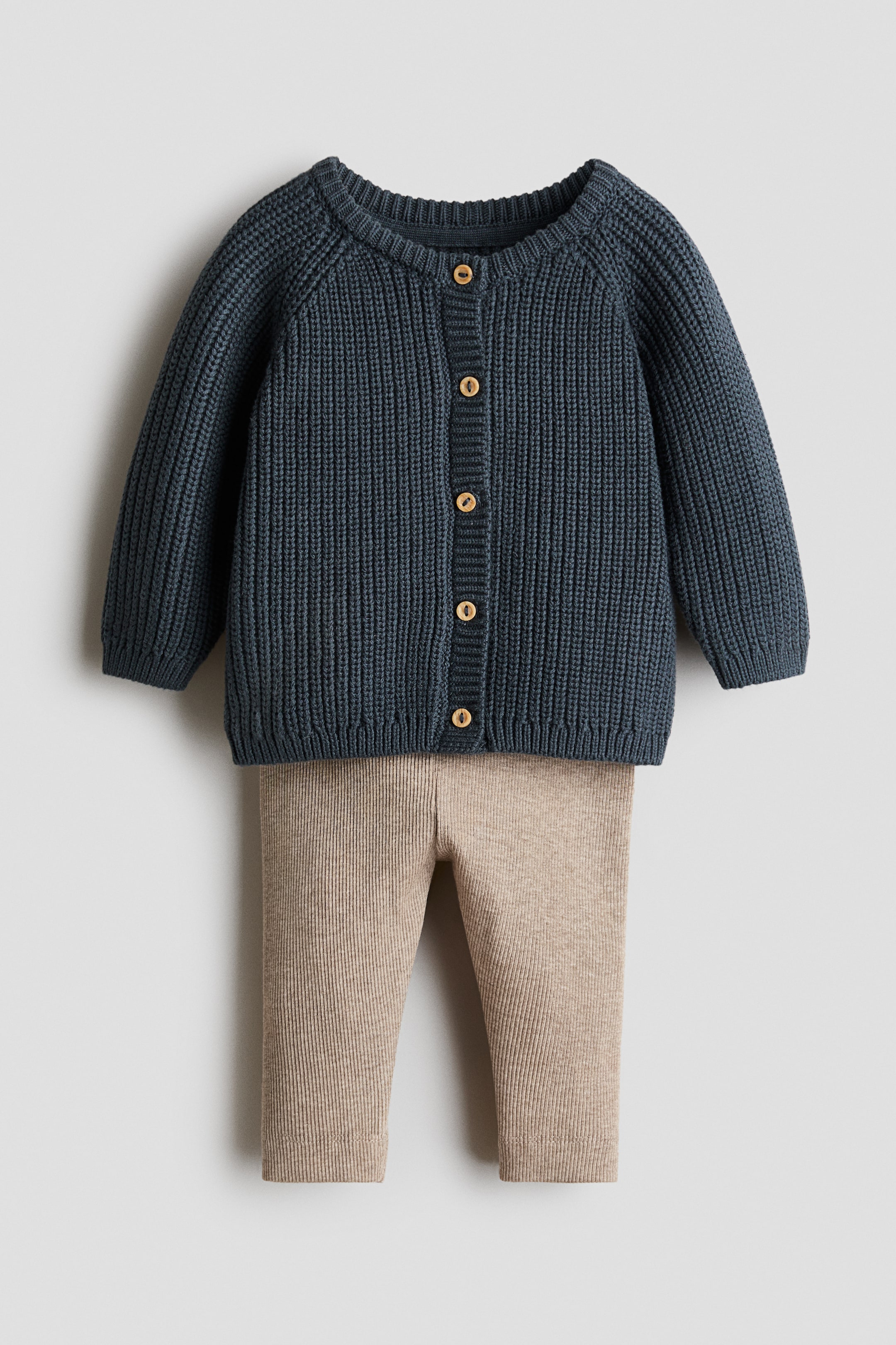 Xem ảnh lớn hơn: Một bộ trang phục trẻ em hai món gồm áo cardigan dệt kim gân dày màu xanh navy đậm và quần dệt kim gân màu be nhạt. Áo cardigan có cổ tròn, tay raglan và hàng khuy cài phía trước với sáu nút màu nâu nhạt.