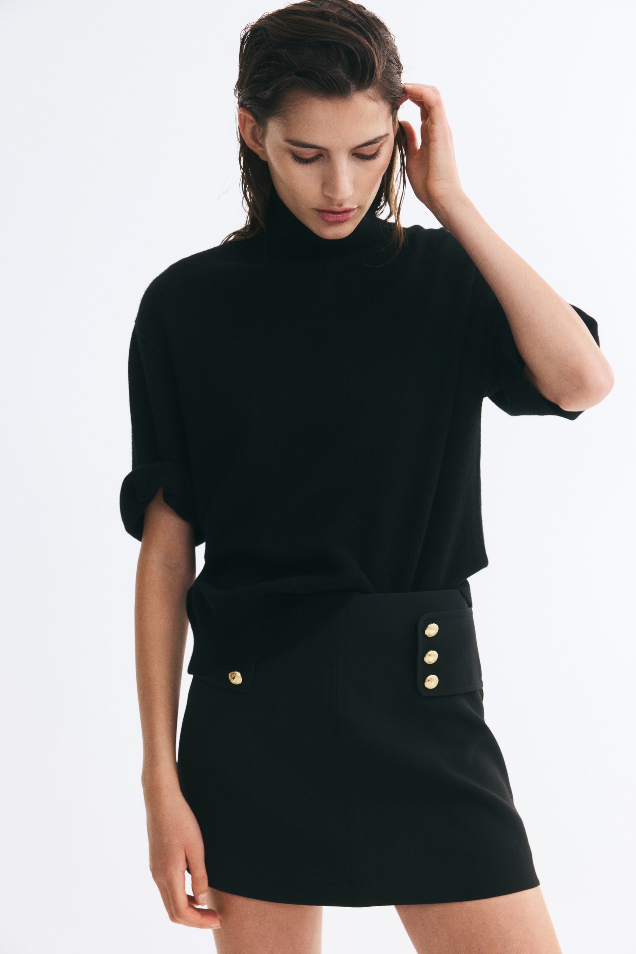 Ampliar la imagen: La modelo lleva una falda-pantalón corta negra con cintura alta, confeccionada con amplias solapas laterales adornadas con botones dorados decorativos y una costura frontal visible. La prenda se combina con un top negro de manga corta y cuello alto, metido por dentro.