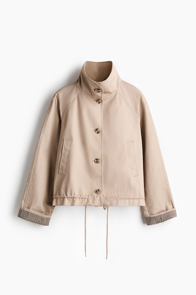 A beige jacket is showcasing its front view, featuring a standing collar, a button-front fastening with brown buttons, and two flap pockets. It is designed with raglan sleeves, rolled cuffs revealing a checkered lining, and a drawstring hem with visible ties. - 1