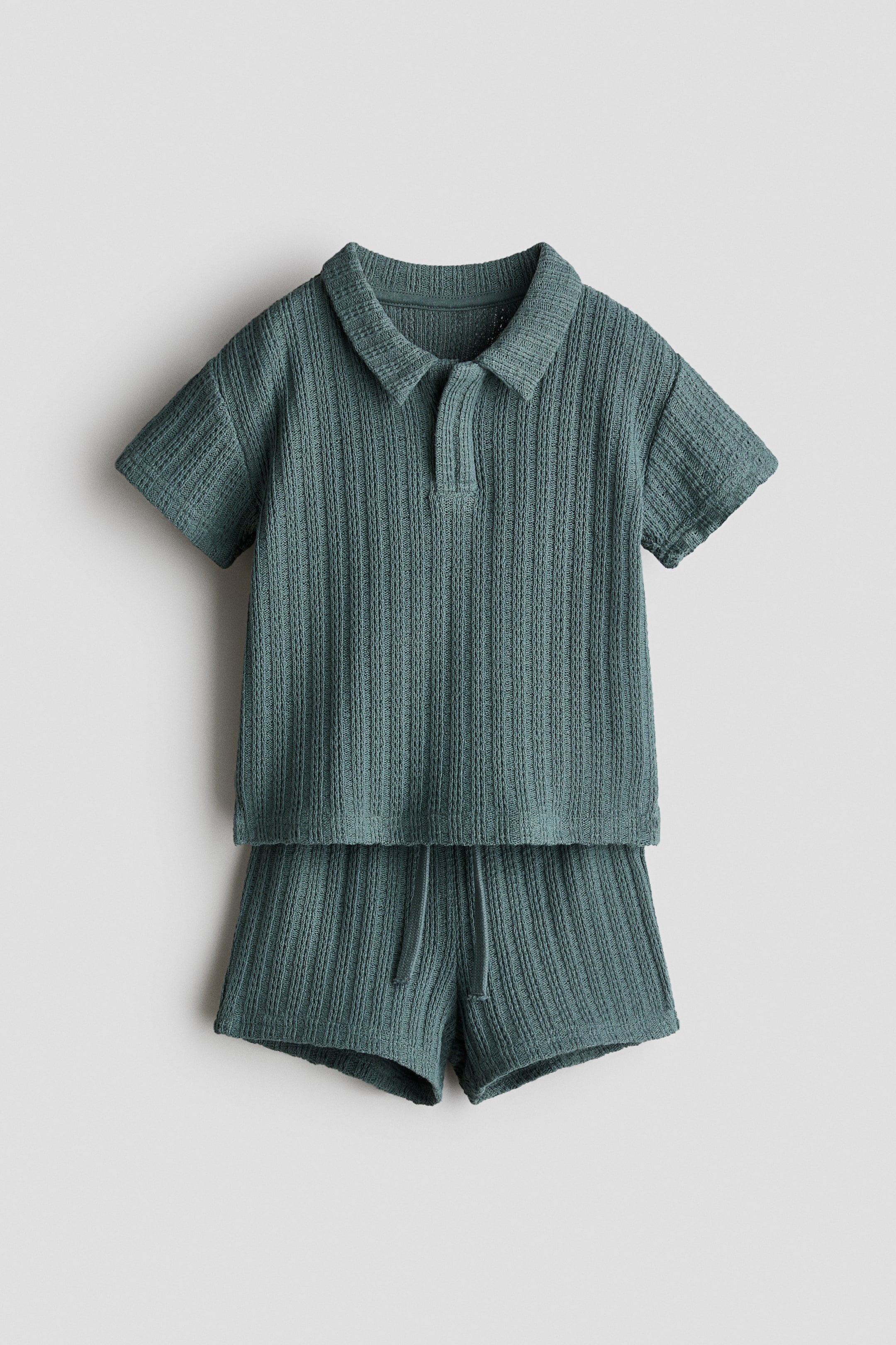 View larger image: A set, presented FRONT, features a teal polo shirt and matching shorts, both crafted from a soft pointelle knit. The polo shirt displays a collar with a V-shaped opening, dropped shoulders, and small slits at the hem, while the shorts showcase an elasticated, drawstring waist.