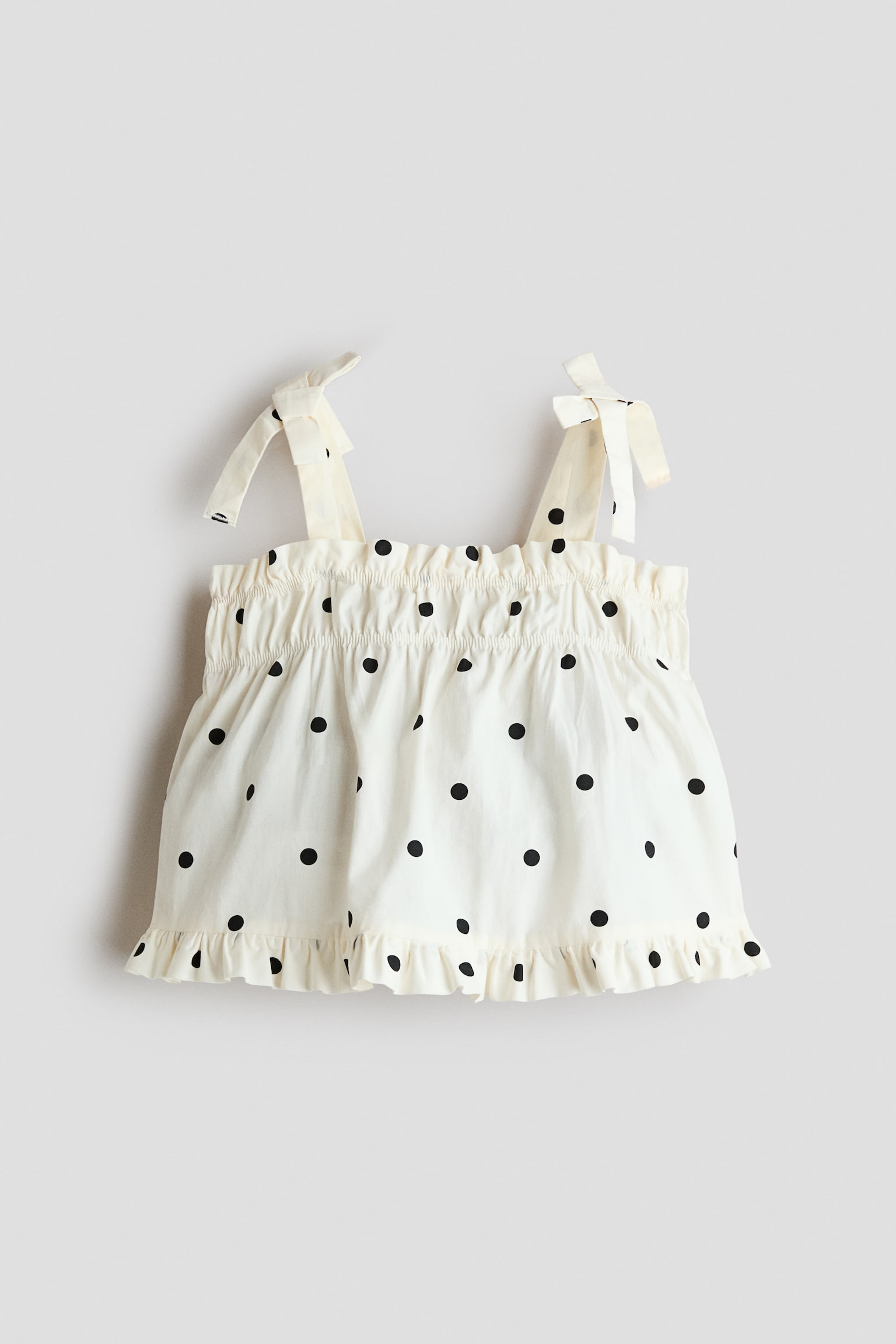 A cream-coloured sleeveless top with black polka dots is facing the front, showcasing self-tie shoulder straps and a smocked, ruffled neckline. The lightweight fabric drapes into a flared silhouette that ends with a ruffled hem.