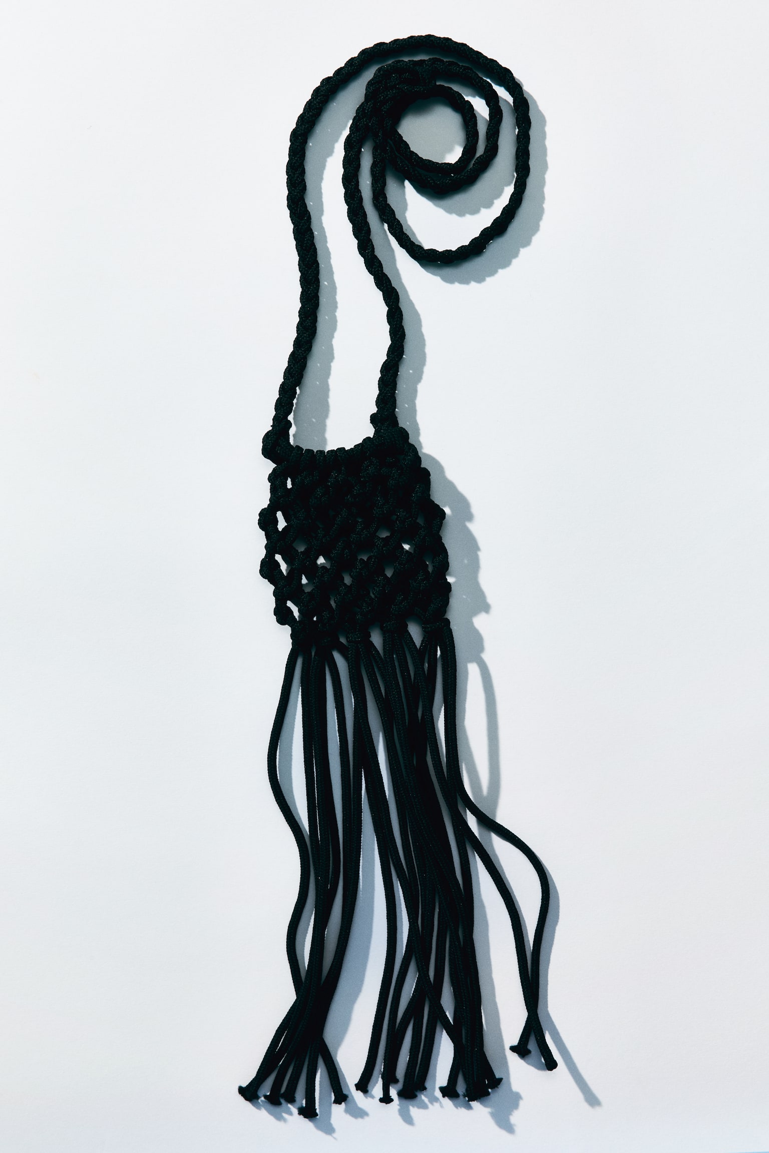 Un accesoriu macrame negru prezintă o curea lungă împletită care formează o buclă în partea de sus, ducând la o secțiune rotundă de macrame cu noduri complexe, din care atârnă mai multe fire de cordon, creând un ciucure. - 1