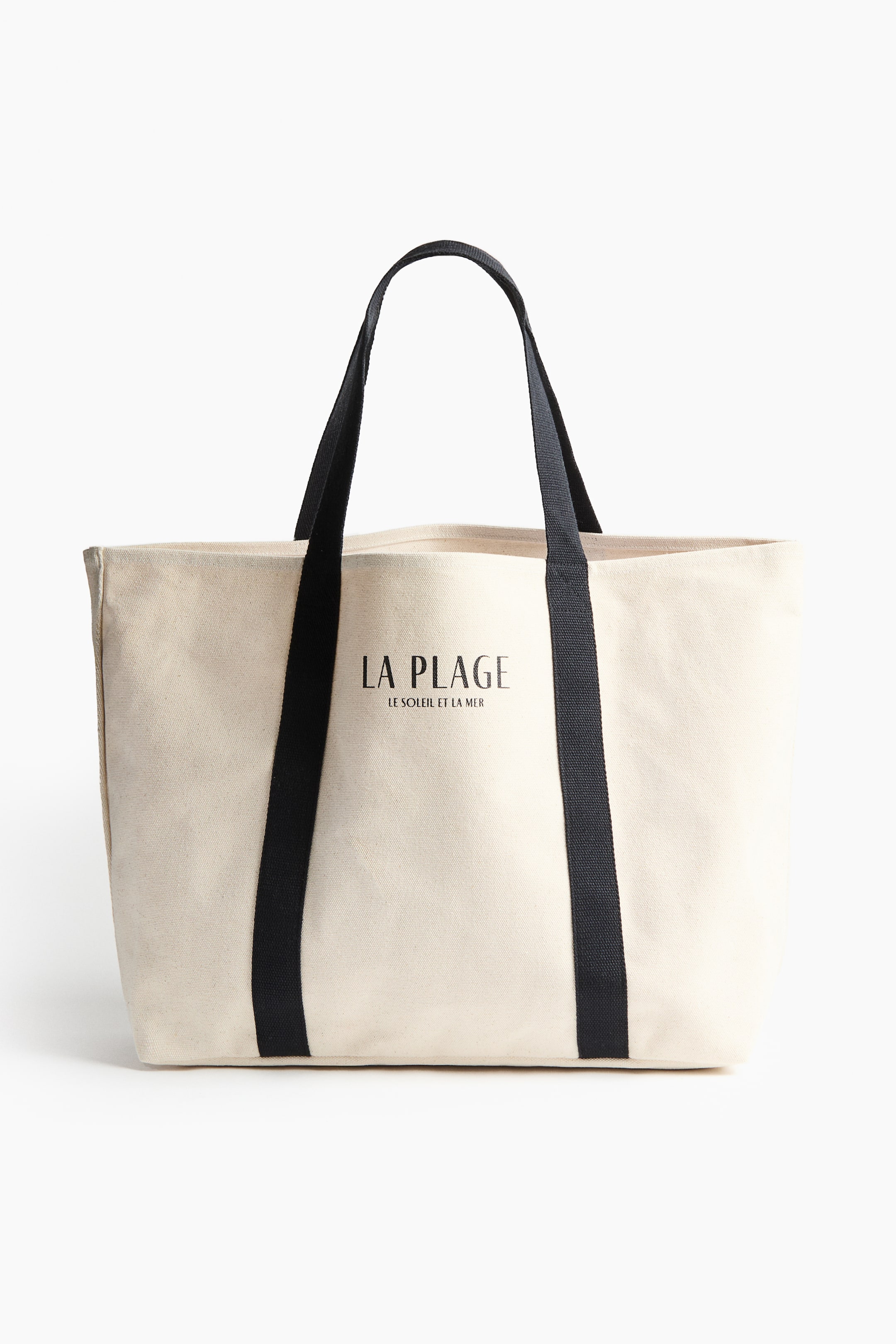 拡大画像を表示: クリーム色のキャンバス製トートバッグが正面を見せており、黒い布製ショルダーストラップが2本付いています。バッグには大きな黒い文字で「LA PLAGE」とプリントされ、その下に小さめの黒い文字で「LE SOLEIL ET LA MER」と印字されています。