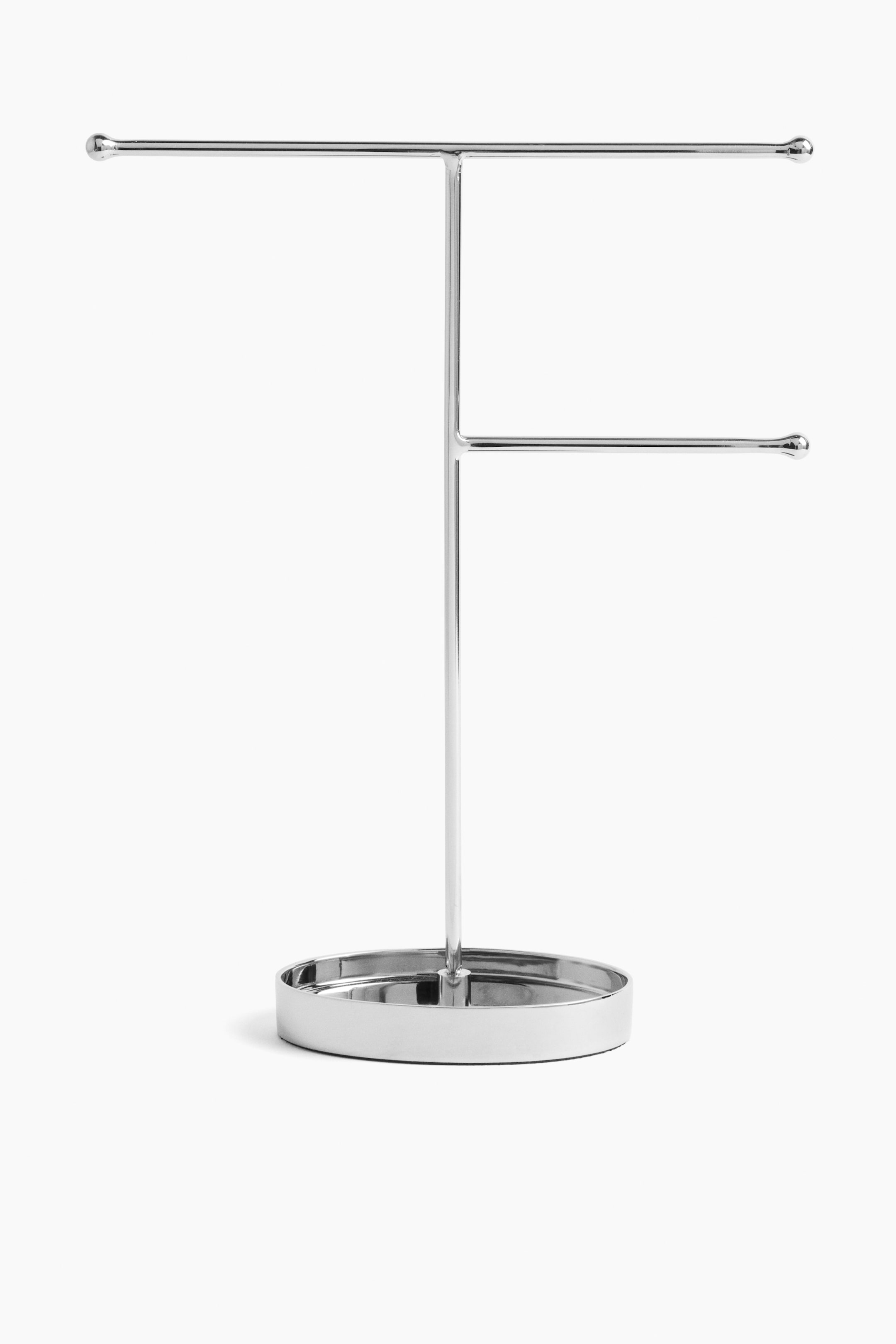 View larger image: A silver-toned metal jewellery stand is standing upright, featuring a circular tray base with a central vertical rod. The vertical rod supports a long horizontal hanging bar at the top and a shorter horizontal hanging bar lower down on one side, both finished with rounded ends.