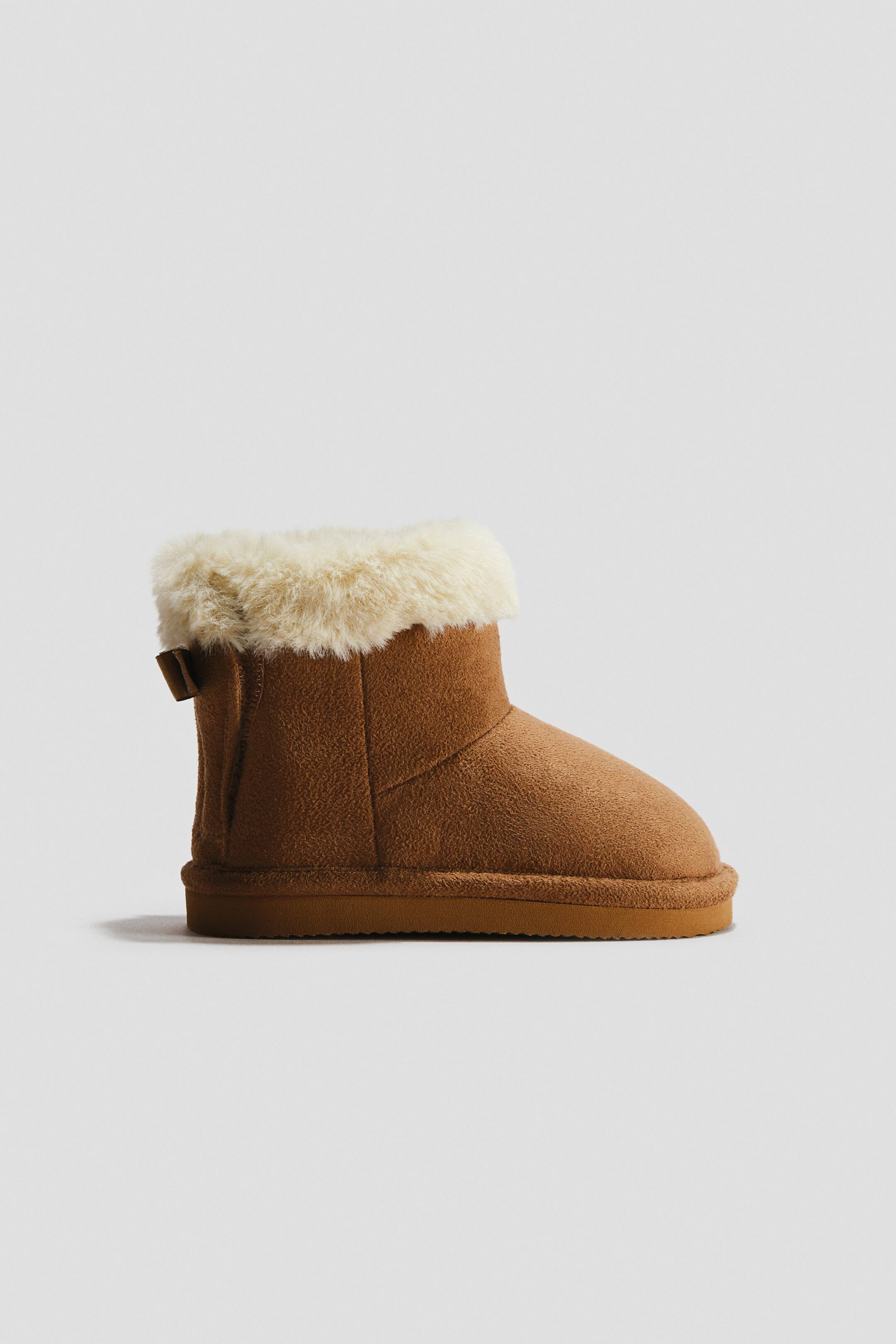 View larger image: A brown boot is facing to the right, featuring a soft, fleece-like exterior with a cream-coloured faux fur lining visibly spilling over the top edge. A small brown pull tab is positioned at the back of the shaft, and a flat, textured brown sole is supporting it.