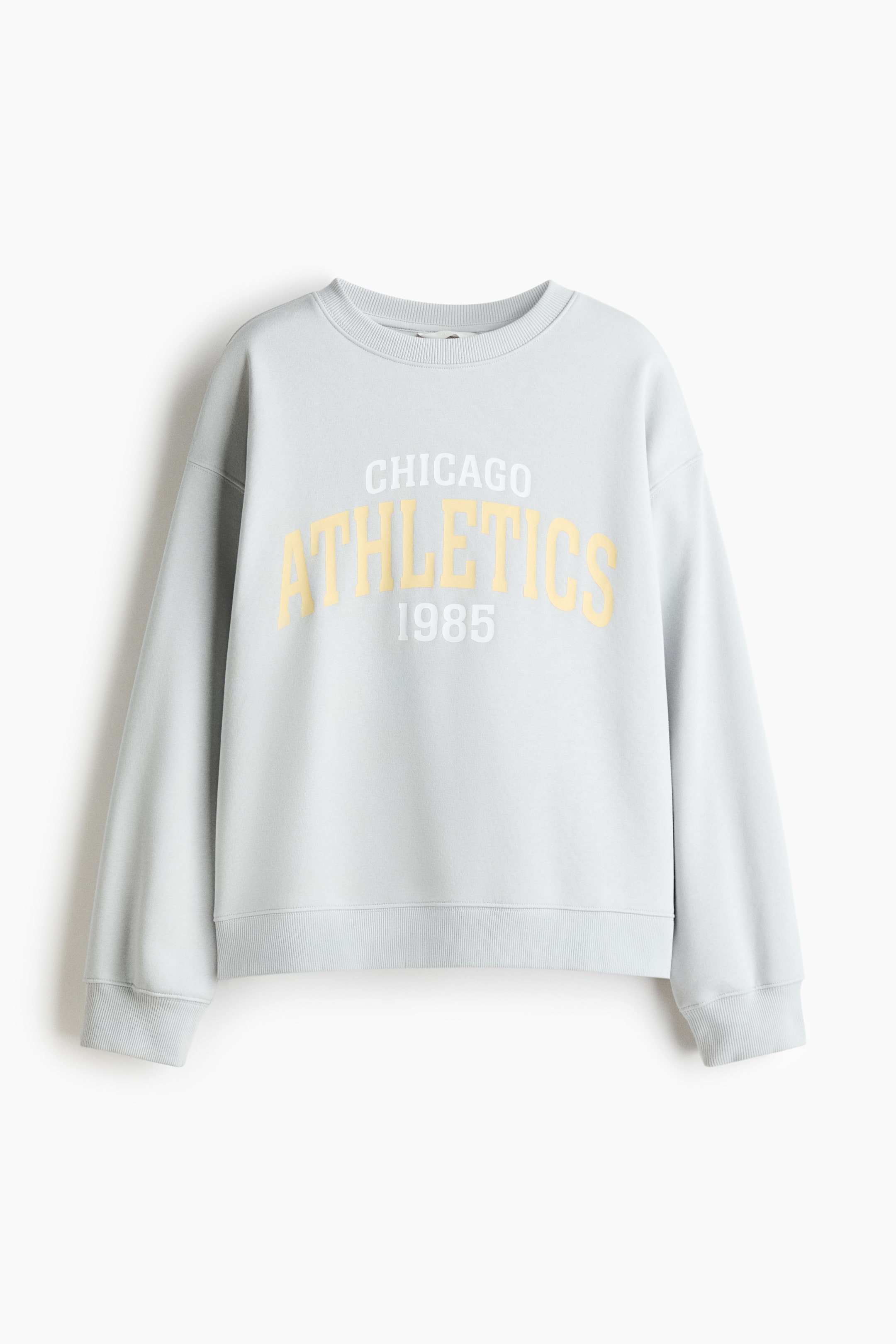 View larger image: An oversized light blue sweatshirt is shown from the FRONT, featuring a "CHICAGO ATHLETICS 1985" text motif in white and yellow on its chest. It is designed with a round, rib-trimmed neckline, dropped shoulders, long sleeves, and ribbed cuffs and hem.