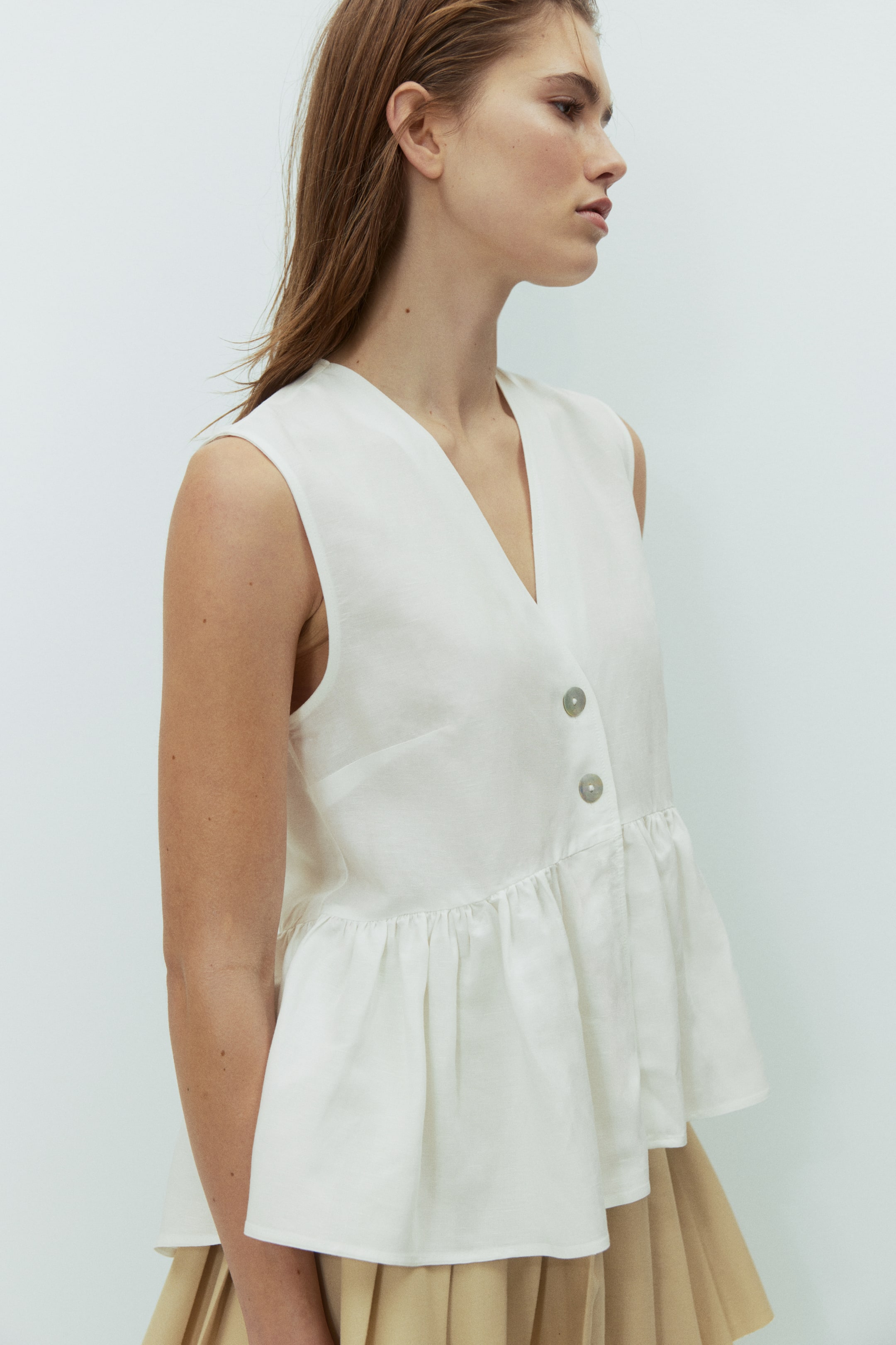 View larger image: A loose-fit, sleeveless white top with a deep V-neckline and two front buttons is worn by the model. It features a gathered seam at the waist that creates a flared peplum and layers over a light brown pleated skirt.