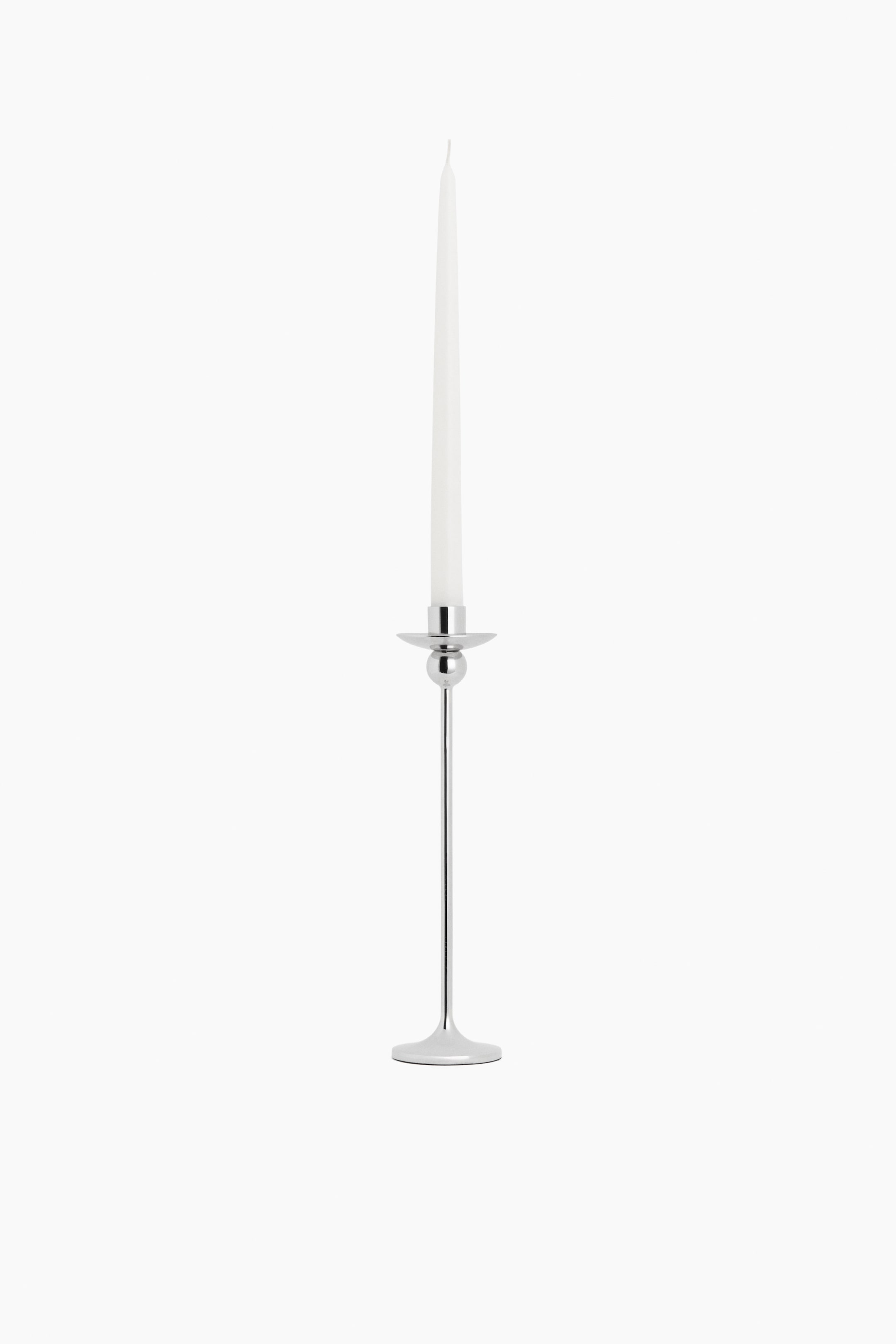 View larger image: A polished silver-toned metal candlestick holds a white taper candle. The candlestick showcases a slender stem, a flat round base, and a small spherical detail below the candle cup.