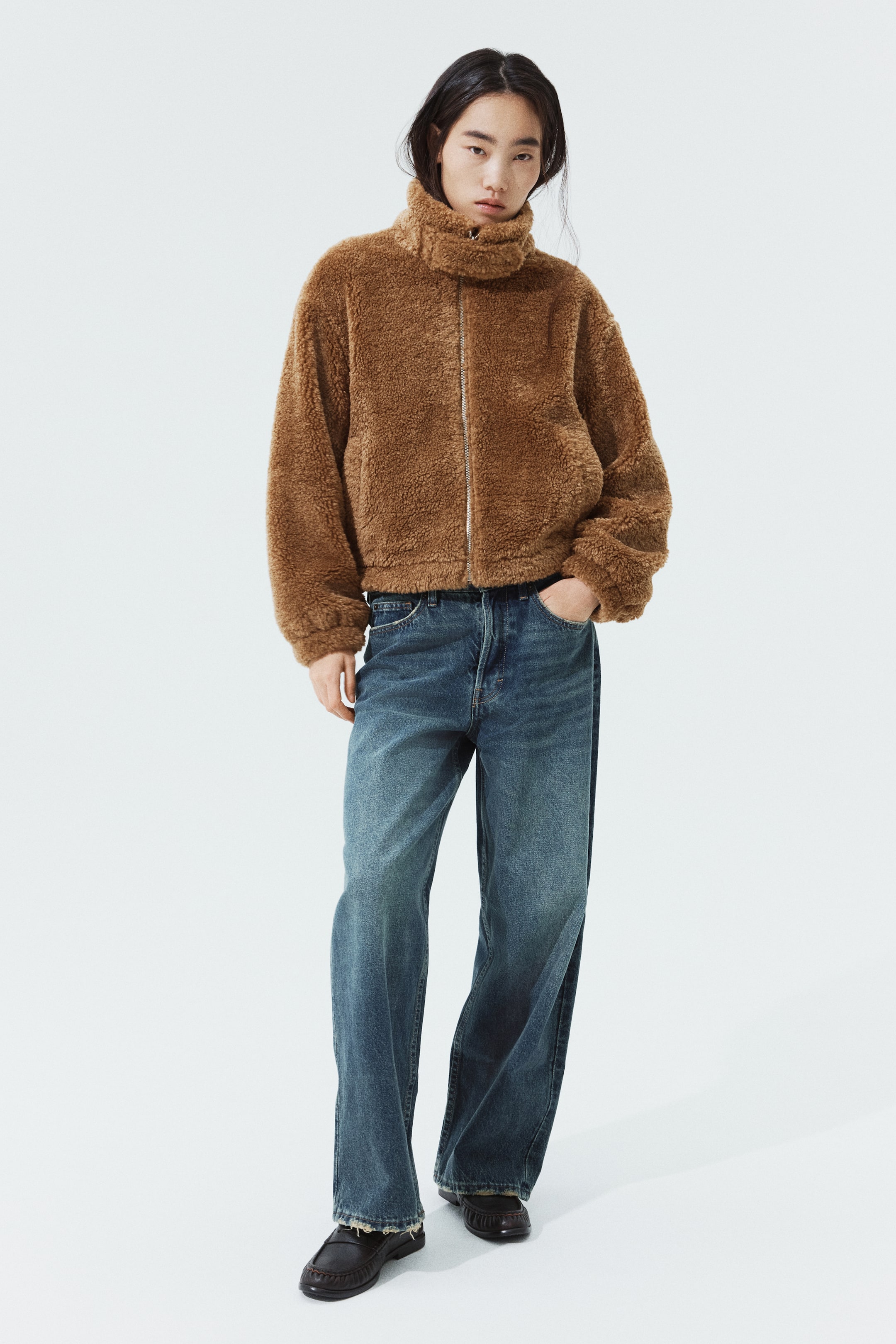 拡大画像を表示: モデルは、スロートラッチ付きの高めのファネルカラー、フロントジッパー、ドロップショルダーが特徴のブラウンのパイルジャケットを着用しています。ジャケットは、ブルーのワイドレッグジーンズとダークカラーのローファーでスタイリングされています。