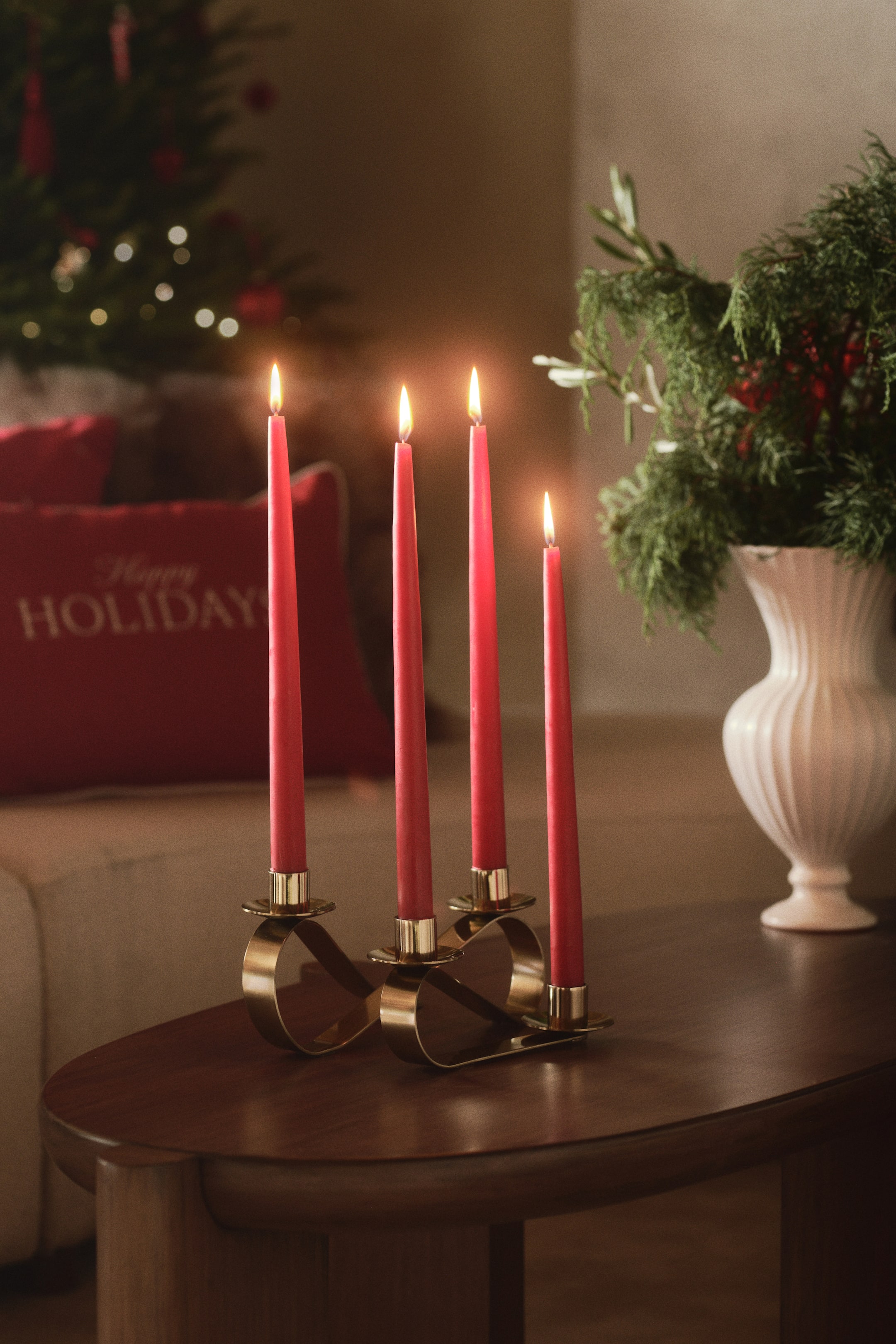 View larger image: A gold-toned stainless steel candelabrum featuring a softly curved, ribbon-inspired design holds four lit red taper candles. The undulating metal band loops to form the base and supports the four cylindrical candle holders at staggered heights.