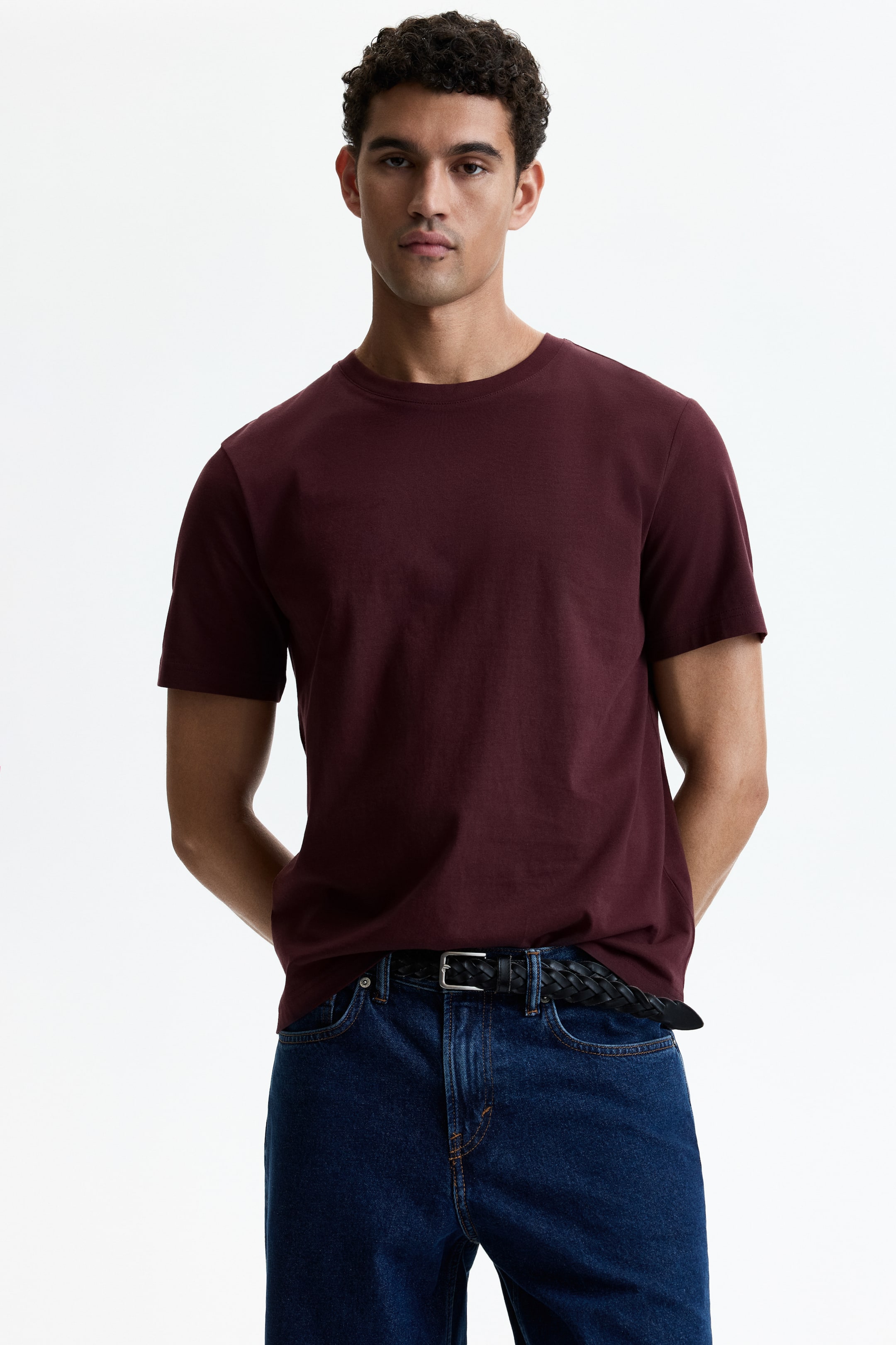 View larger image: The model is wearing a deep red T-shirt, which showcases a round, ribbed neckline, short sleeves, and a straight hemline. The T-shirt is neatly tucked into dark blue jeans, accentuated by a black braided belt at the waist.