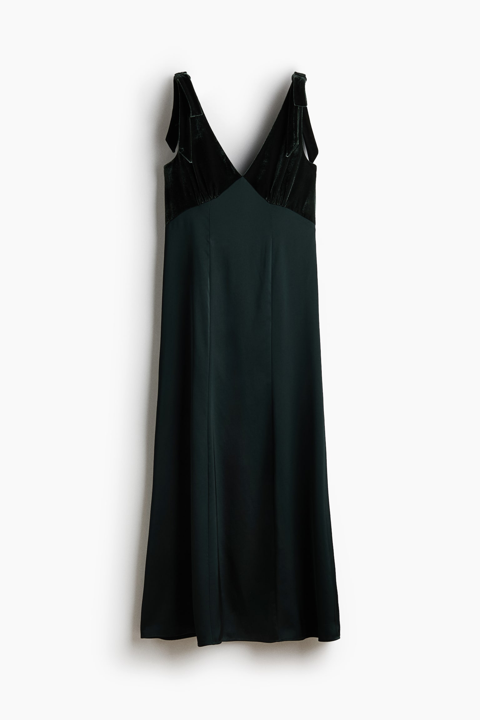 A long, dark green dress is displayed front-facing, featuring a fitted velvet bodice with a deep V-neckline, gathered cups, and wide shoulder straps adorned with decorative bows. The dress flows into a softly draping satin A-line skirt with visible vertical seams running down the front.
