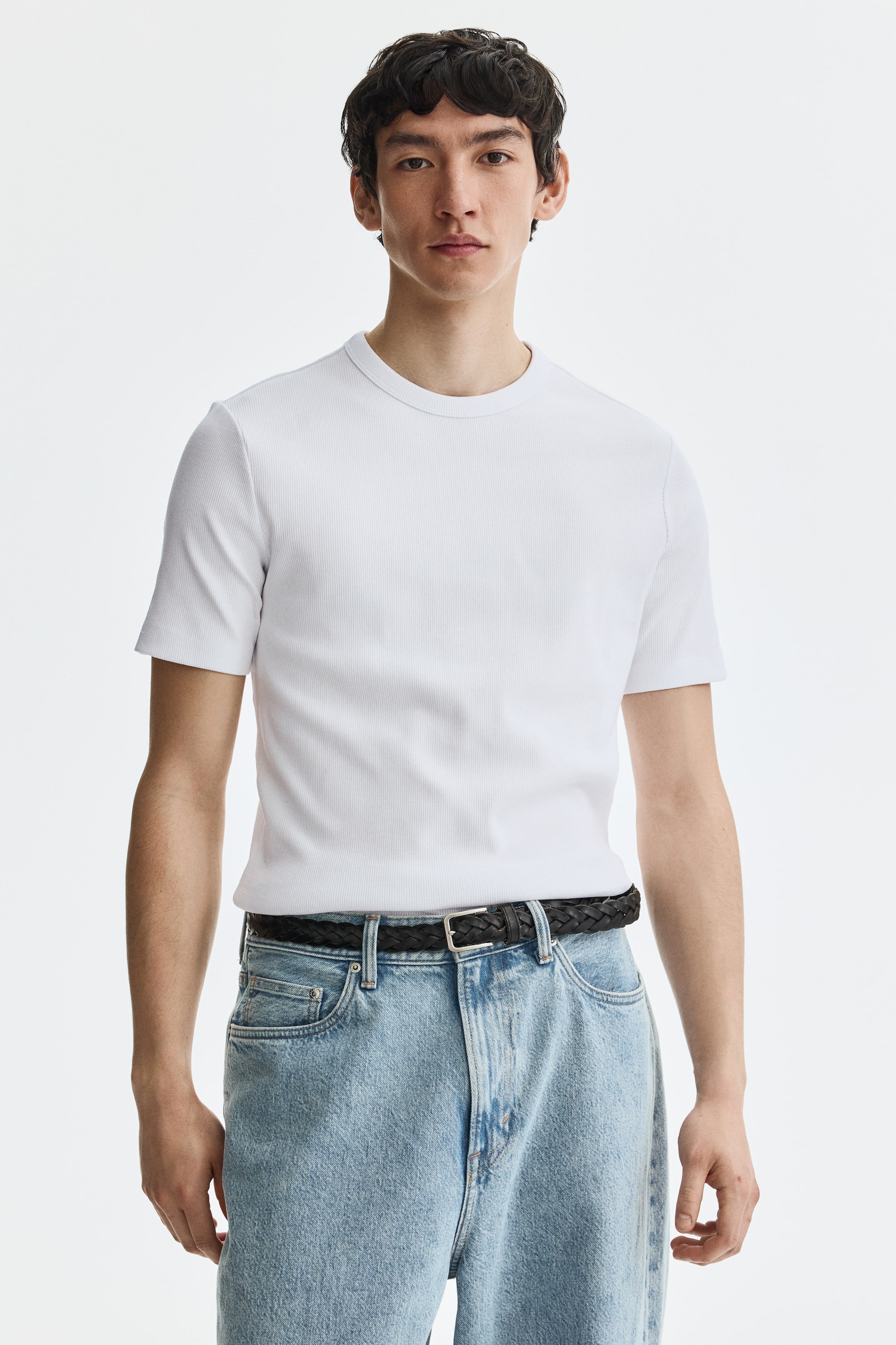 View larger image: A white, finely ribbed crew neck t-shirt is displayed from a front-facing angle. This short-sleeved t-shirt fits snugly and is tucked into light-wash jeans, secured by a black braided belt.