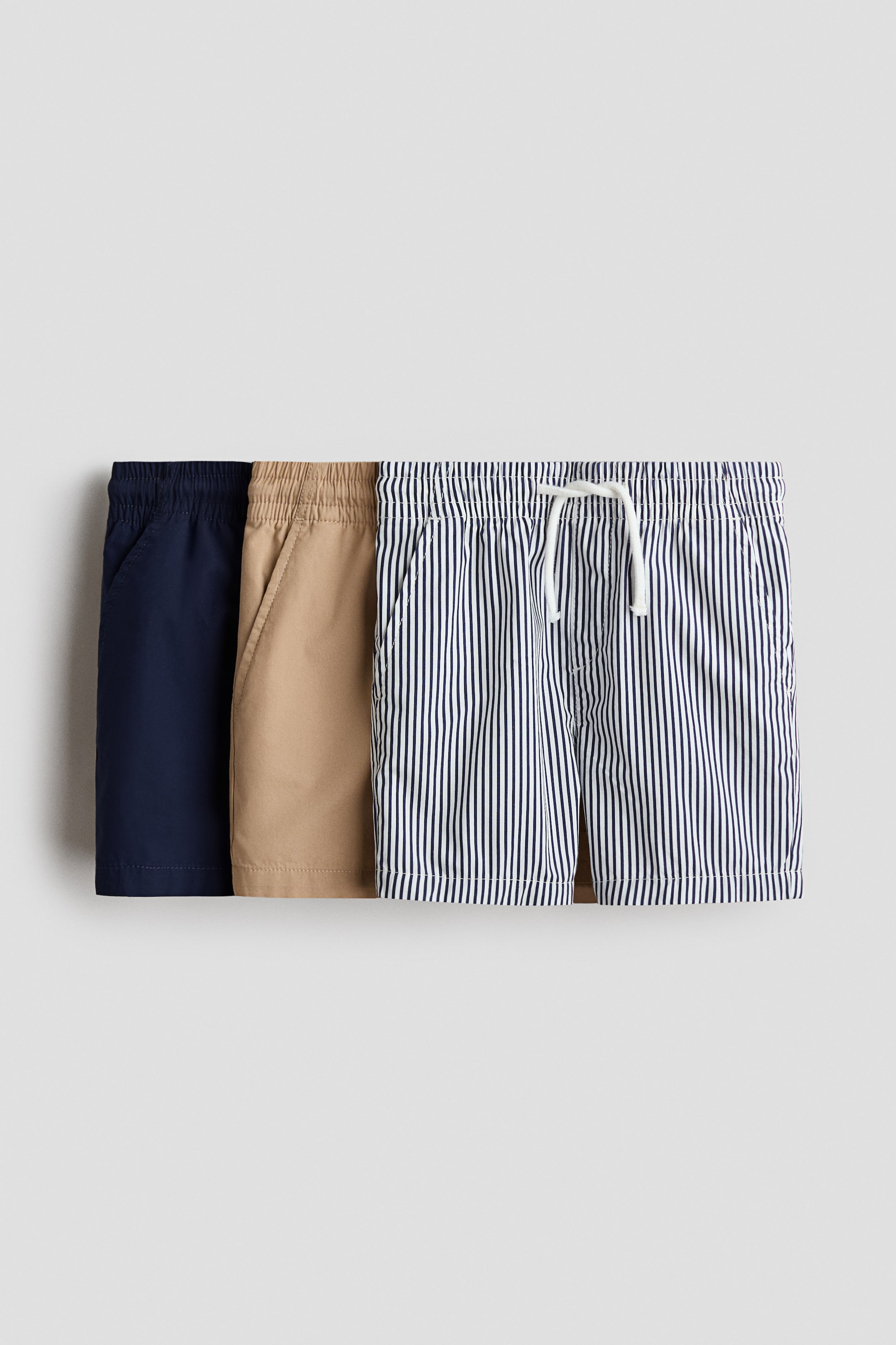 View larger image: Three pairs of crisp cotton poplin shorts are showcased from the front, each featuring an elasticated, drawstring waist, a faux fly, and diagonal side pockets. One pair is solid navy blue, another is solid khaki, and the third pair is white with thin navy vertical stripes.