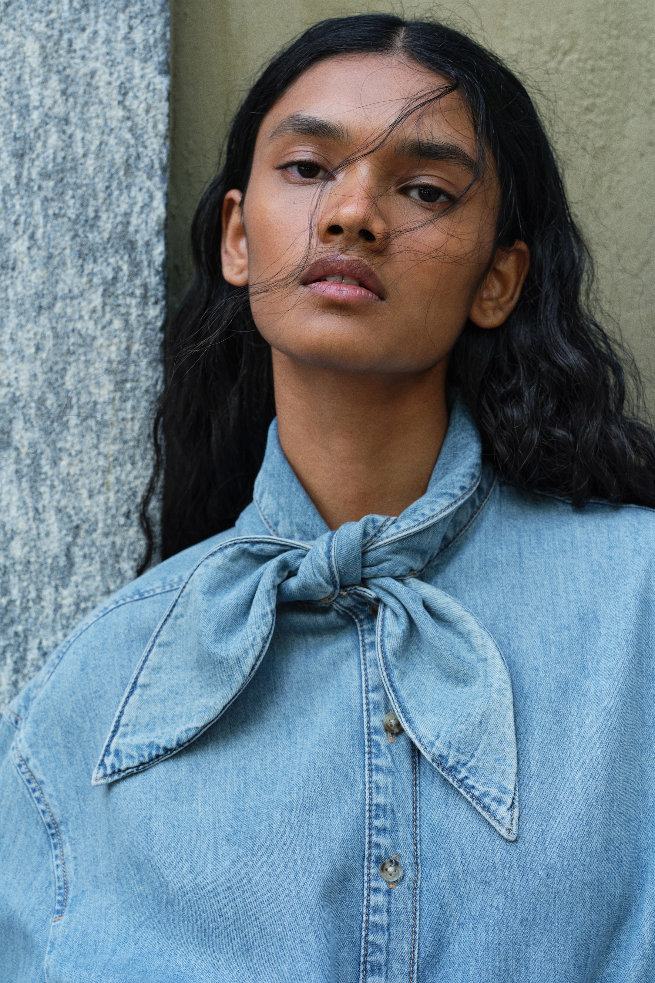 Größeres Bild ansehen: Das Model trägt ein hellblaues Denimhemd mit einem schalartigen Kragen, der am Hals zu einem Knoten gebunden ist. Das Hemd verfügt über eine durchgehende Knopfleiste und überschnittene Schultern.