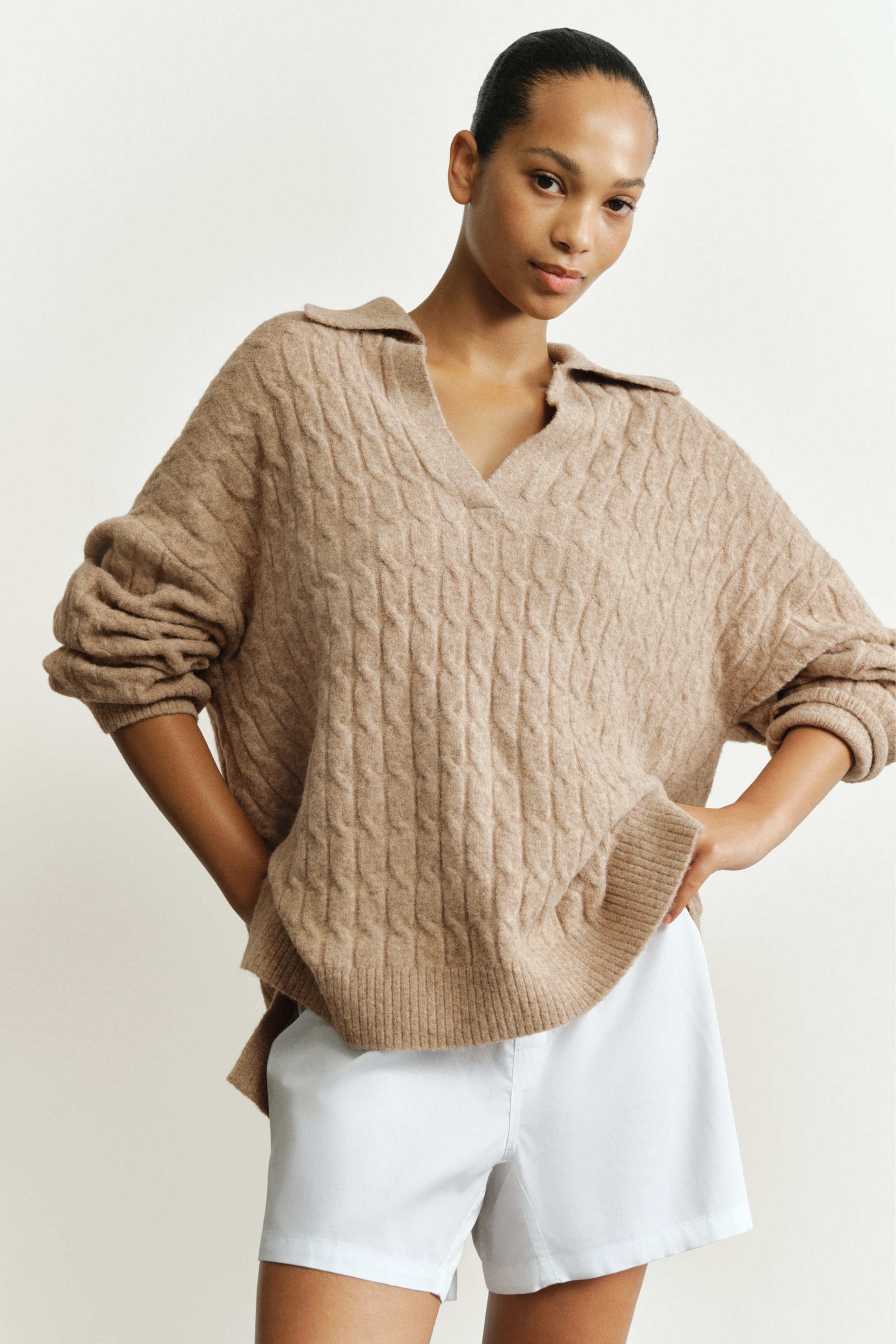 Größeres Bild anzeigen: Ein oversized beiger Zopfpullover wird vom Model getragen und verfügt über einen gerippten offenen Kragen, überschnittene Schultern sowie einen gerippten Saum mit Seitenschlitzen. Der Pullover wird mit weißen Shorts gestylt, während das Model die Hände in die Hüften stützt.