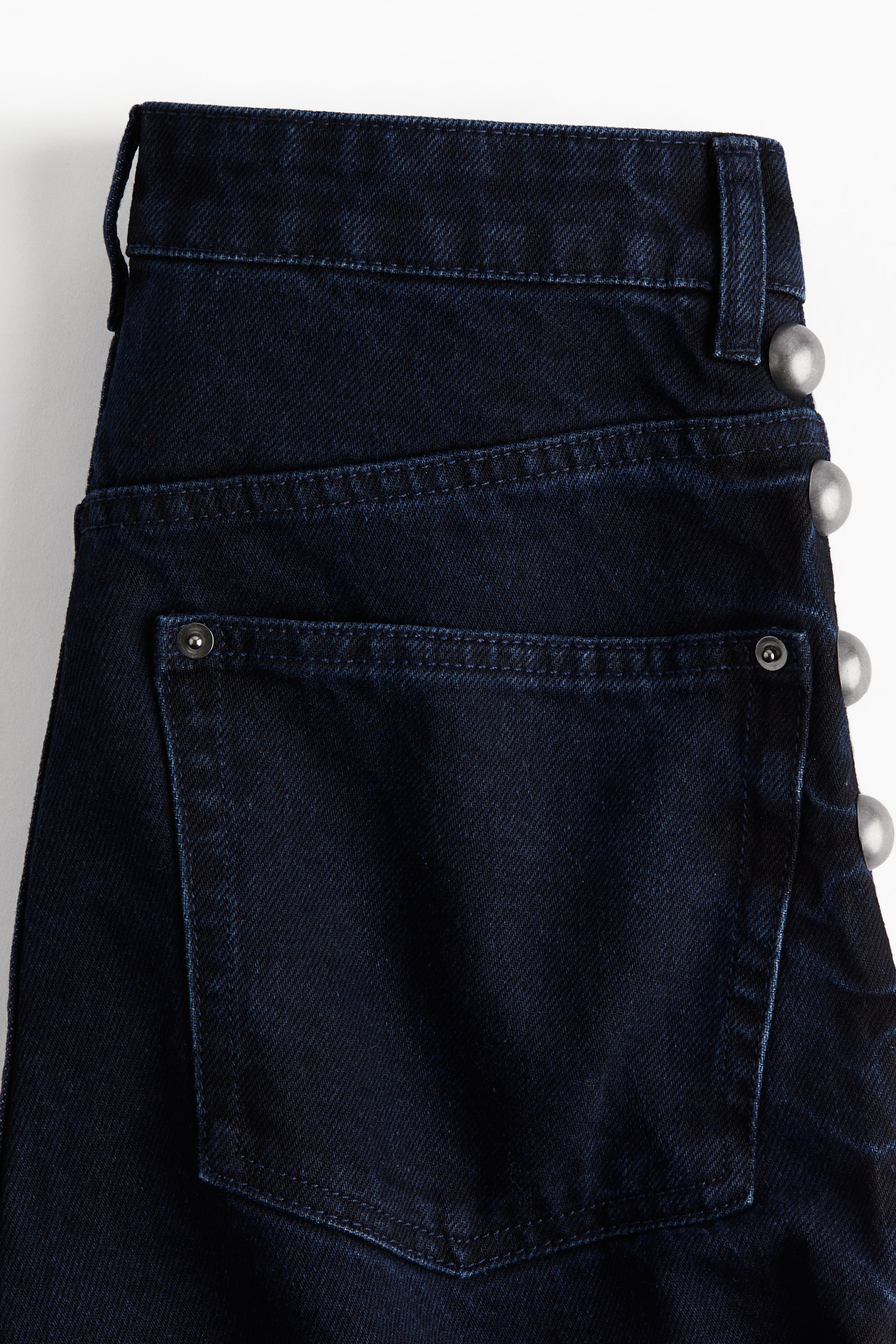 Voir une image plus grande: Un jean en denim de coton rigide bleu foncé est présenté de dos, montrant une taille régulière et une poche arrière. Des clous métalliques en forme de dôme descendent le long du côté extérieur de la jambe droite.