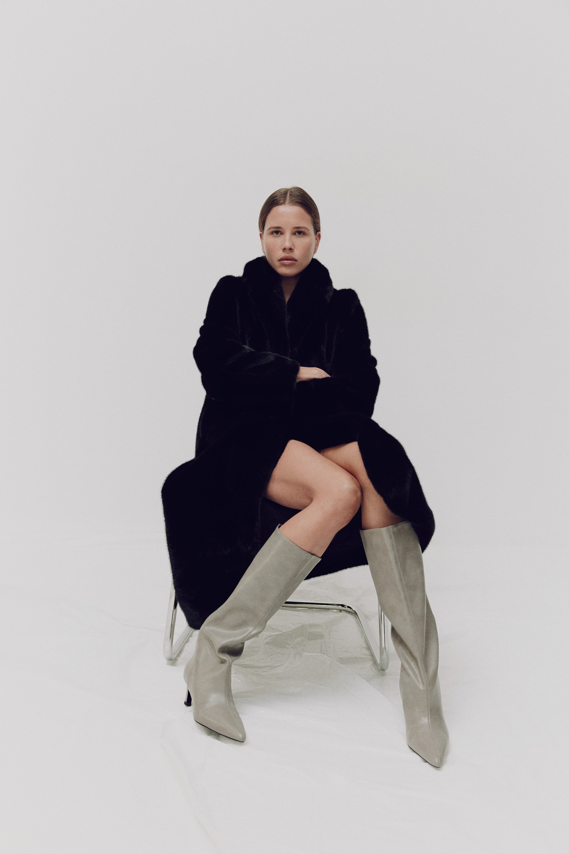 Vezi imaginea mai mare: "Un palton din blană artificială neagră prezintă o textură luxoasă și voluminoasă, mâneci lungi și un guler larg. Este drapat lejer, extinzându-se dincolo de genunchi, cu partea din față deschisă, dezvăluind picioarele purtătorului."