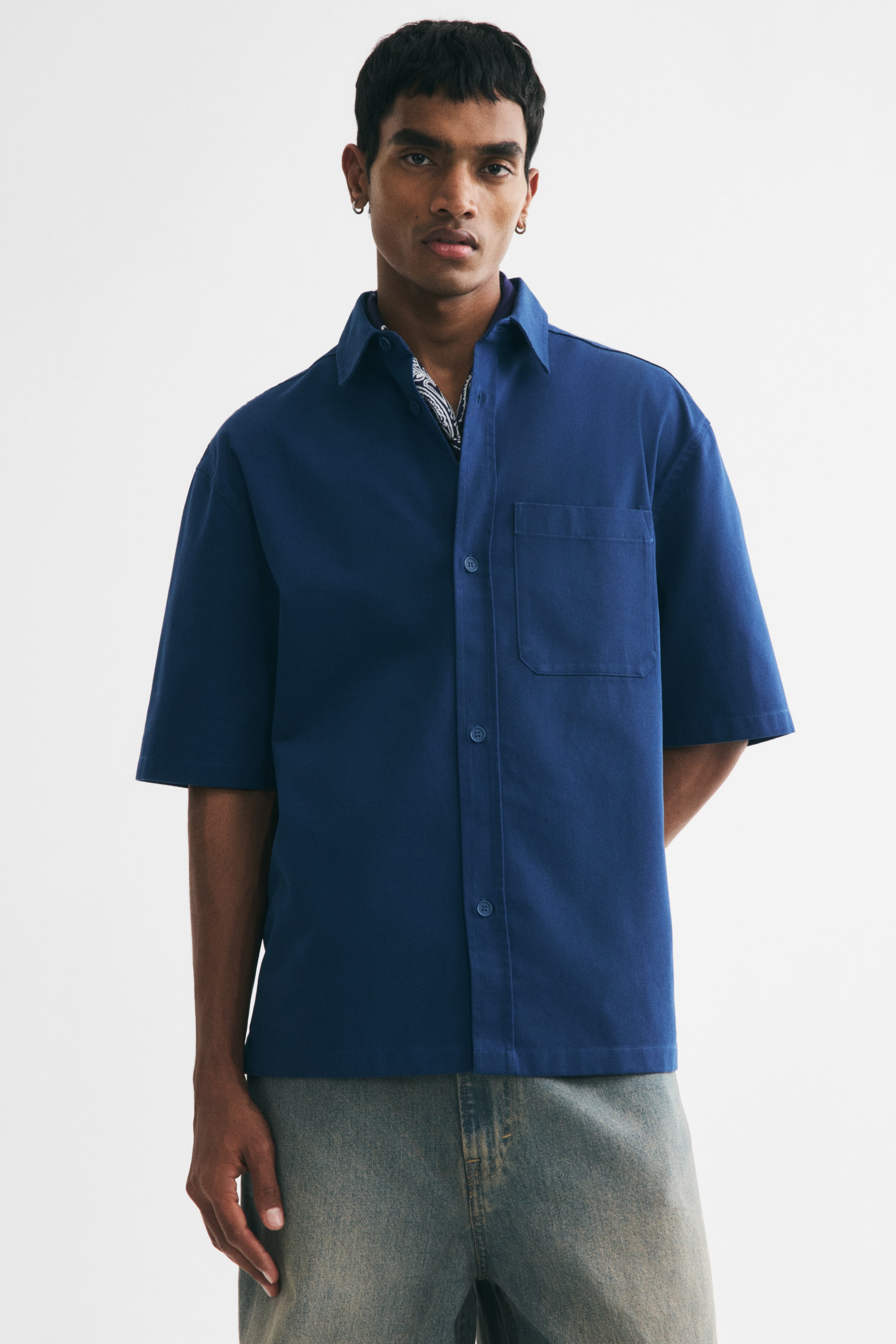 View larger image: The model is wearing a dark blue short-sleeved shirt with a collar, a classic button-front, and an open chest pocket. The shirt boasts a relaxed fit and a straight-cut hem, accented with a patterned bandana at the collar.