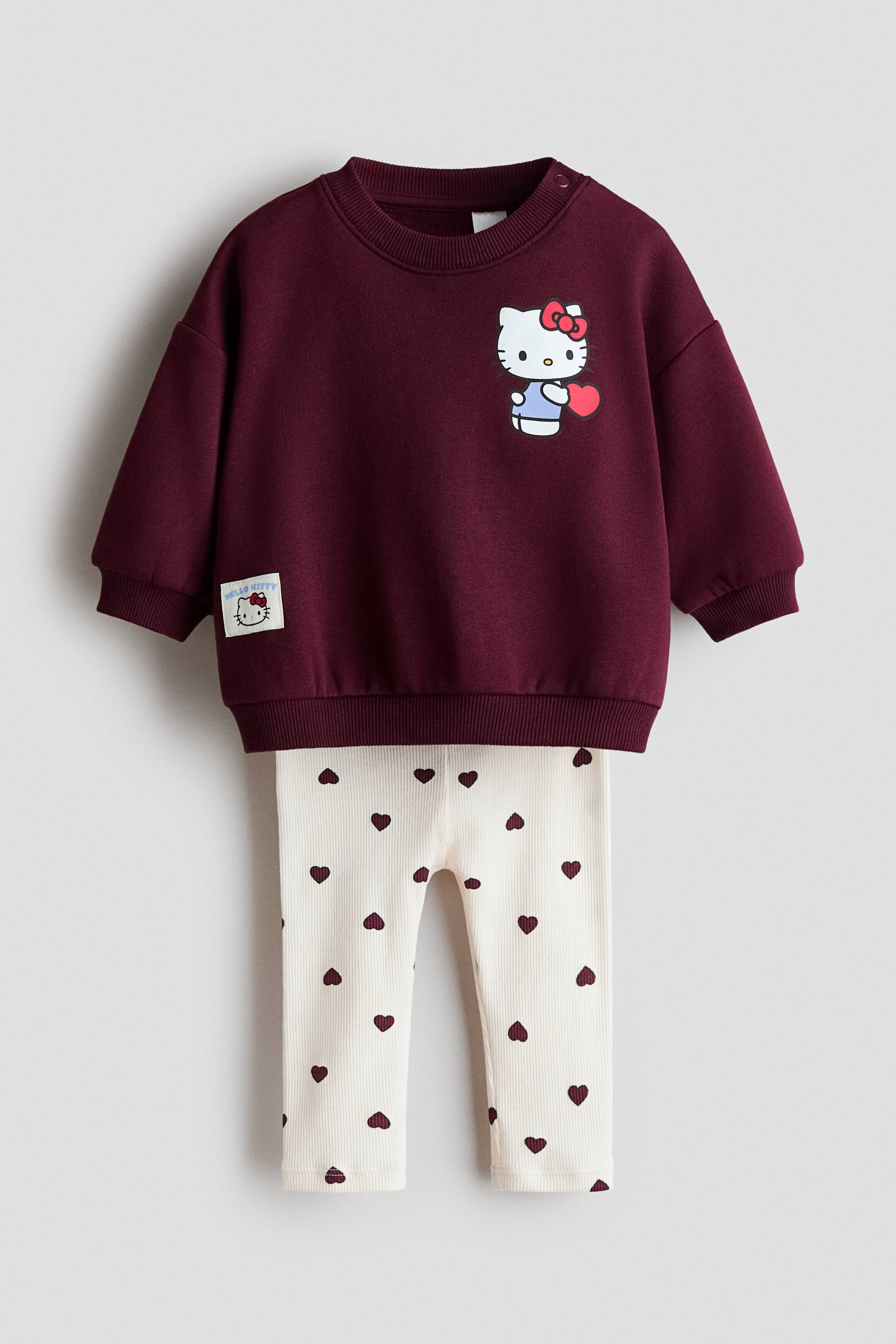 View larger image: The two-piece printed set is shown from the front, featuring a maroon long-sleeved sweatshirt and cream-coloured ribbed leggings. The crew-neck sweatshirt displays a Hello Kitty graphic on the chest, along with ribbed collar, cuffs, and hem detailing, while the matching leggings showcase an all-over pattern of small maroon hearts.