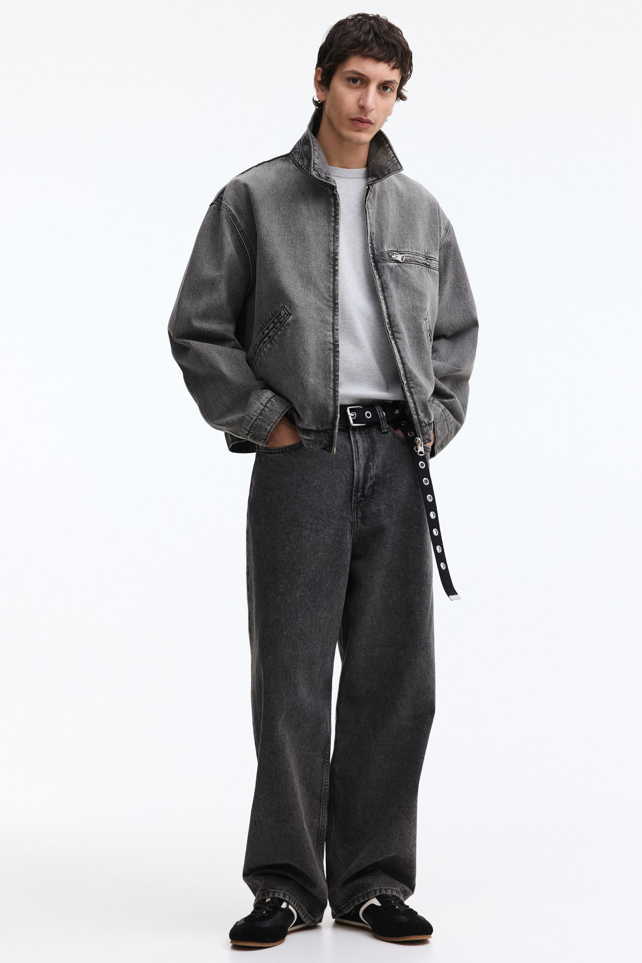 Ingrandisci l'immagine: Una giacca di jeans grigio lavato viene mostrata dal davanti, caratterizzata da un colletto rialzato, una chiusura a zip frontale, due tasche laterali e una tasca sul petto con zip, indossata sopra una t-shirt grigio chiaro. Jeans in denim grigio scuro a vestibilità ampia sono indossati, completati da una cintura nera con occhielli.