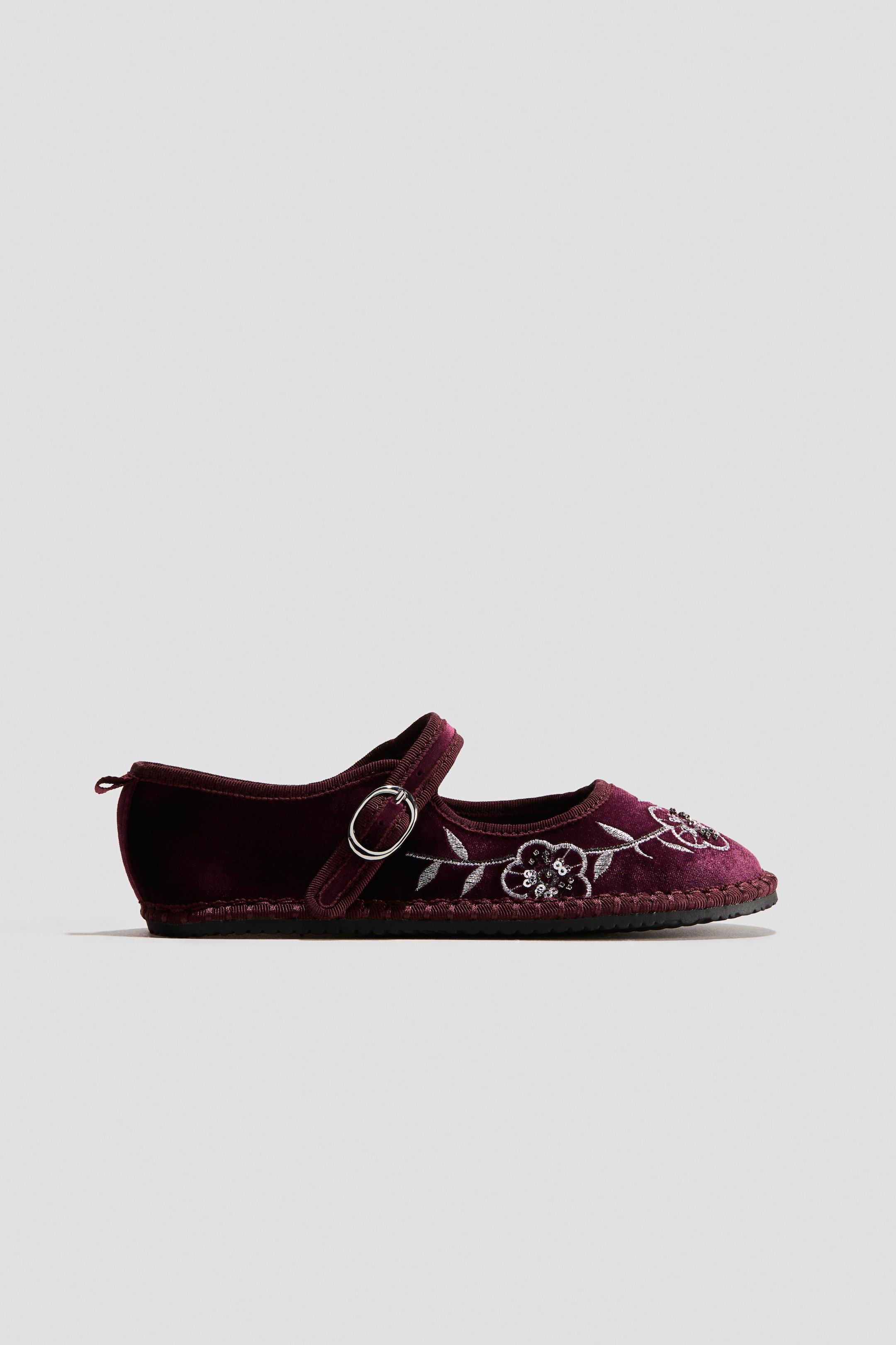 View larger image: A burgundy velour Mary Jane shoe is shown in side profile, featuring silver floral embroidery with beaded details on the toe. The shoe is designed with an adjustable instep strap with a silver metal buckle, a grosgrain-trimmed collar, a heel pull loop, and a slim black sole.